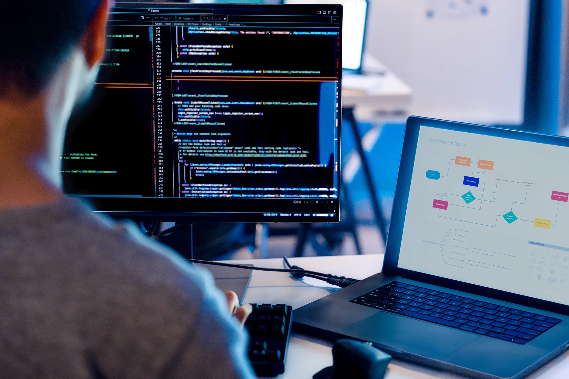 Person working on a computer with code displayed on a monitor and a development flowchart on a laptop screen.