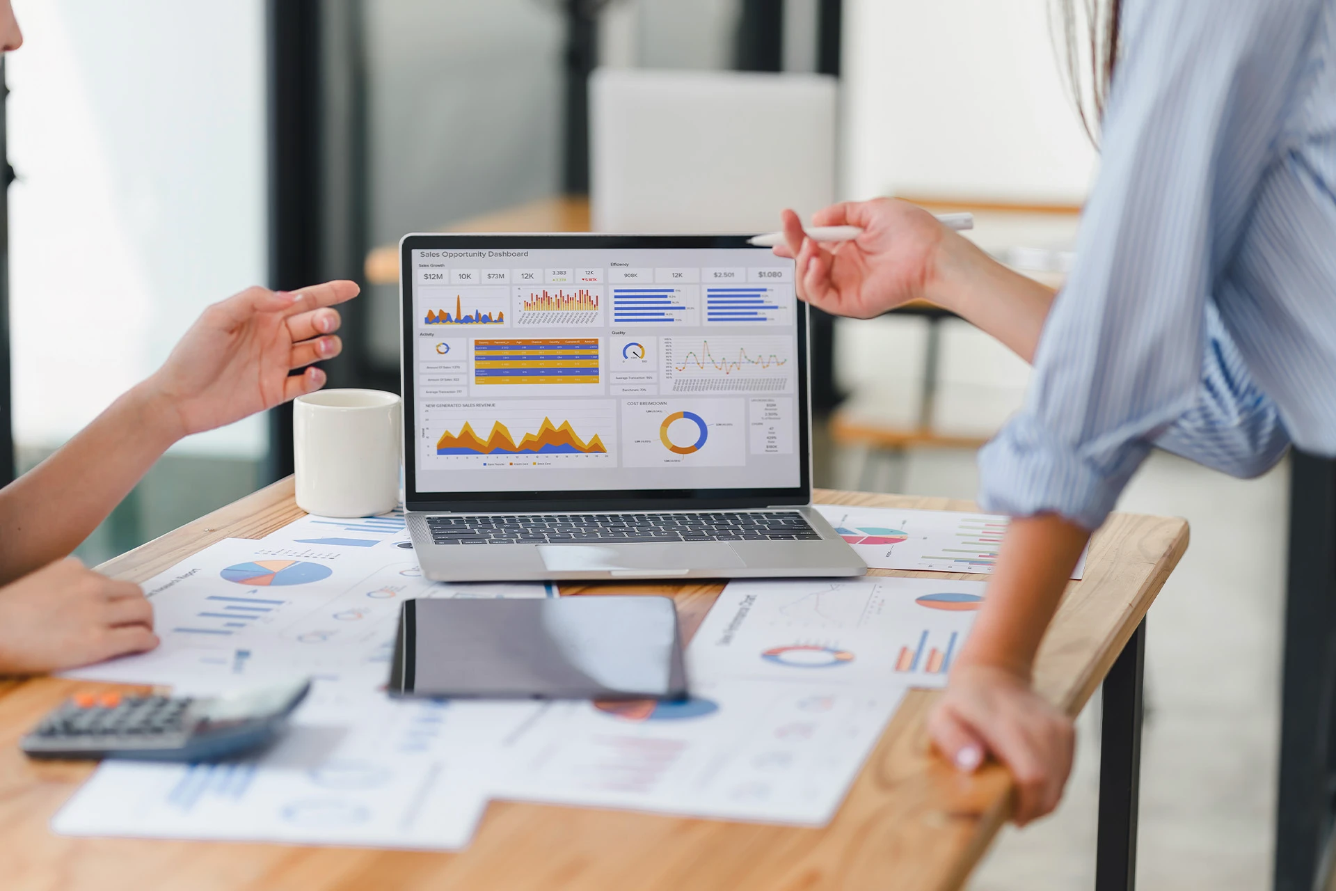 Two people discussing sales dashboard charts and graphs displayed on a laptop at a desk with documents and a tablet.
