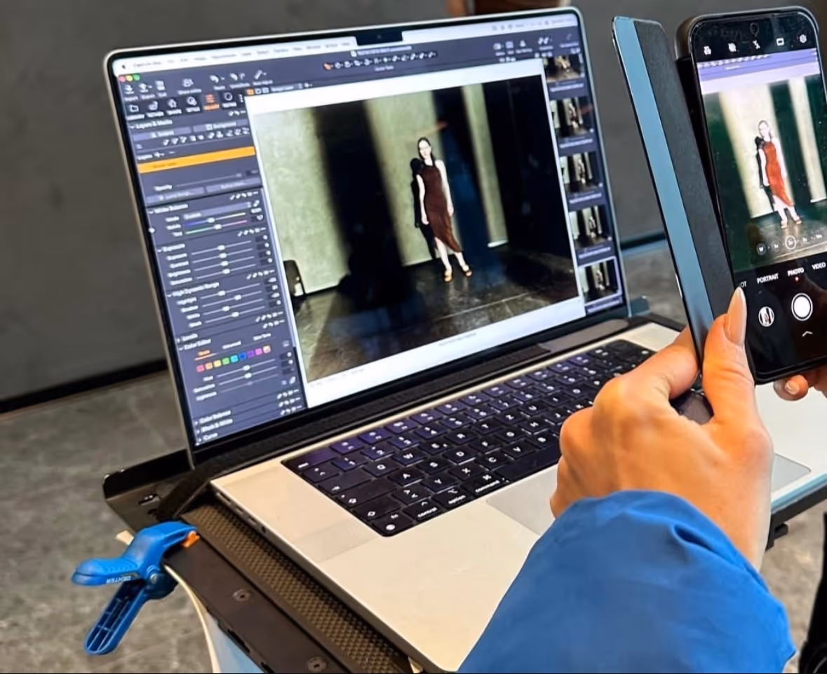 Person in a blue jacket holding a smartphone capturing a photo of a woman in a dark dress on a laptop screen with photo editing software open.