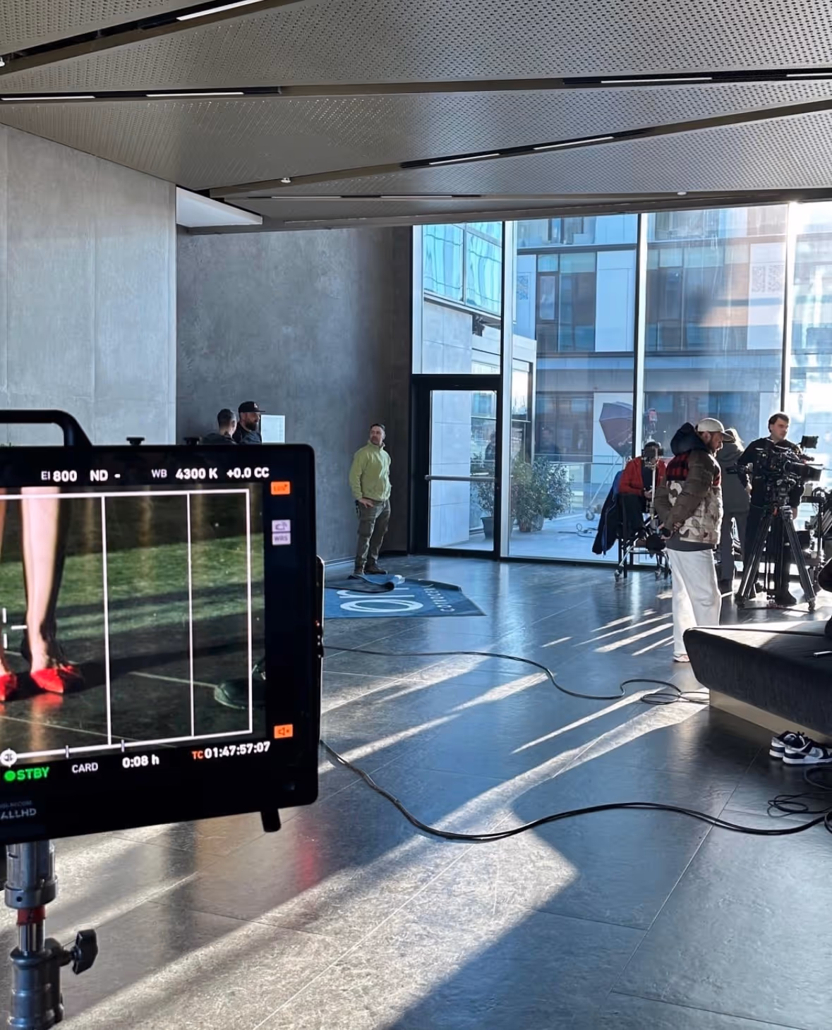 Film set inside a modern building with a camera monitor showing a close-up of red shoes, crew members adjusting equipment, and natural light streaming through large windows.