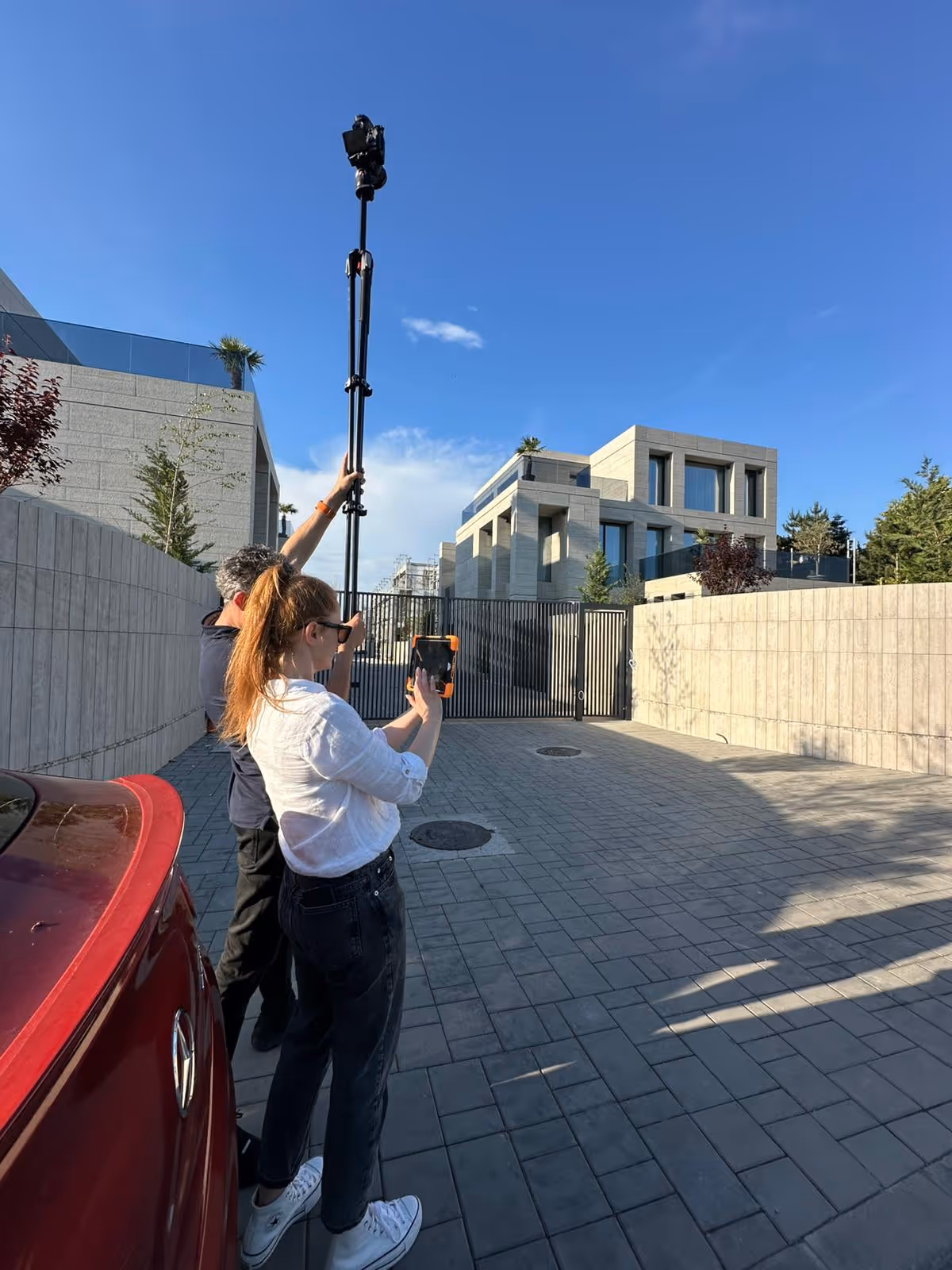 Two people using a tall camera pole and a tablet to photograph or scan a large modern house behind a gated driveway.