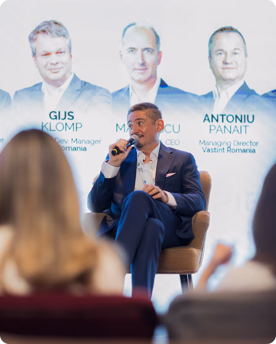 Man in a navy suit speaking into a microphone while seated on stage during a panel discussion.