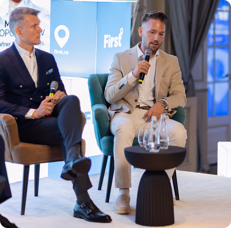 Man in a beige suit sitting on a teal chair holding a microphone, speaking at an indoor event with a table holding water bottles and glasses in front of him.