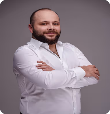 Smiling bearded man wearing a white dress shirt with arms crossed against a gray background.