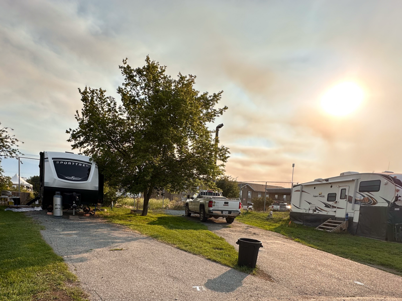 RV park with two parked RVs and a white pickup truck under a cloudy evening sky with the sun shining brightly.