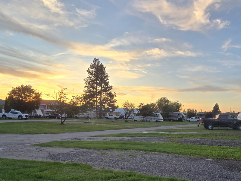 Sunset over an RV park with parked trucks, campers, and tall trees under a partly cloudy sky.