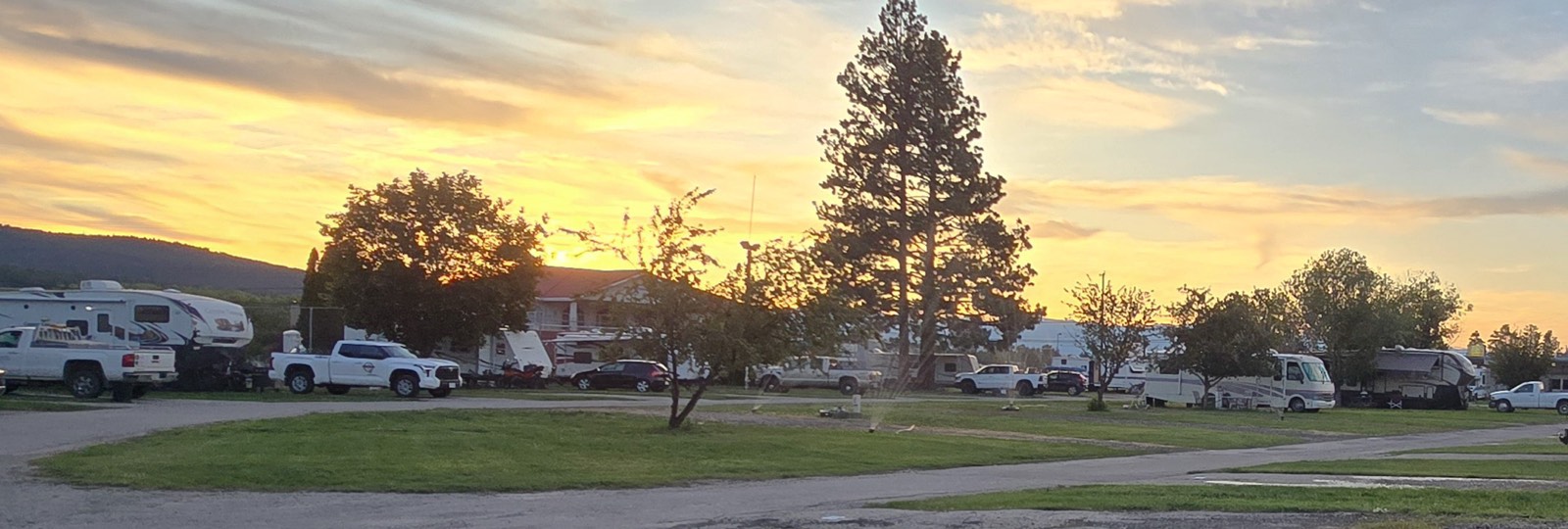 RV park at sunset with parked trucks, trailers, and RVs amid trees and greenery.
