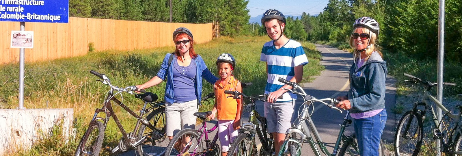 Four people wearing helmets standing with their bicycles on a paved trail surrounded by greenery and a wooden fence.