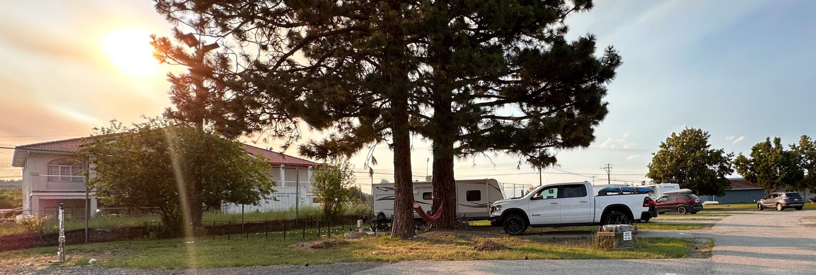 Sunset view of an RV park with a white pickup truck, a camper trailer, two tall pine trees with a red hammock hanging between them, and a gravel road.