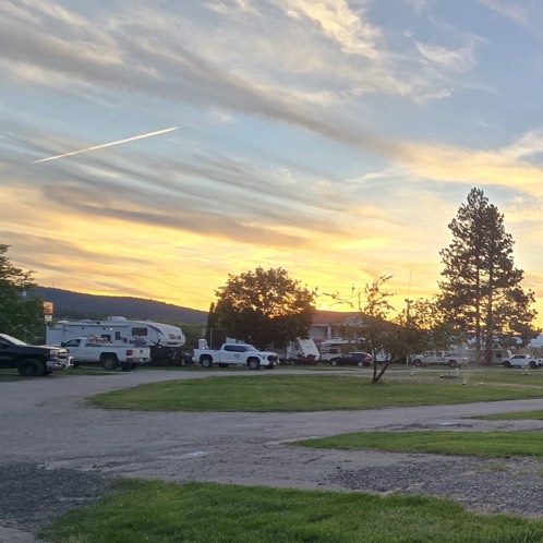 Sunset sky with scattered clouds over a park with RVs, pickup trucks, and trees on grassy and paved ground.