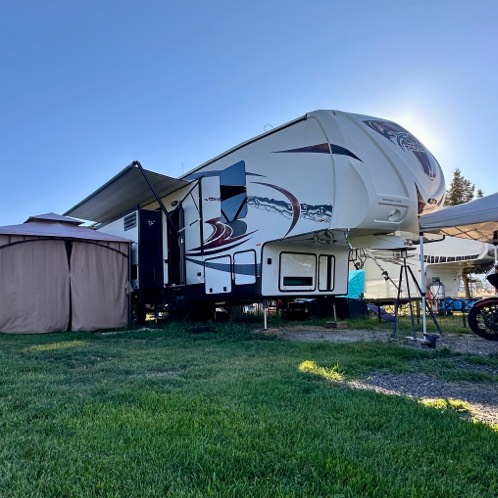 Large white fifth-wheel RV with slide-out and extended awning parked on grass beside a brown tent and other RVs under a clear blue sky.