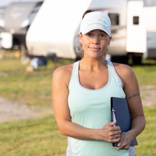 Woman in a light blue tank top and white cap holding a clipboard, standing outdoors near RVs.