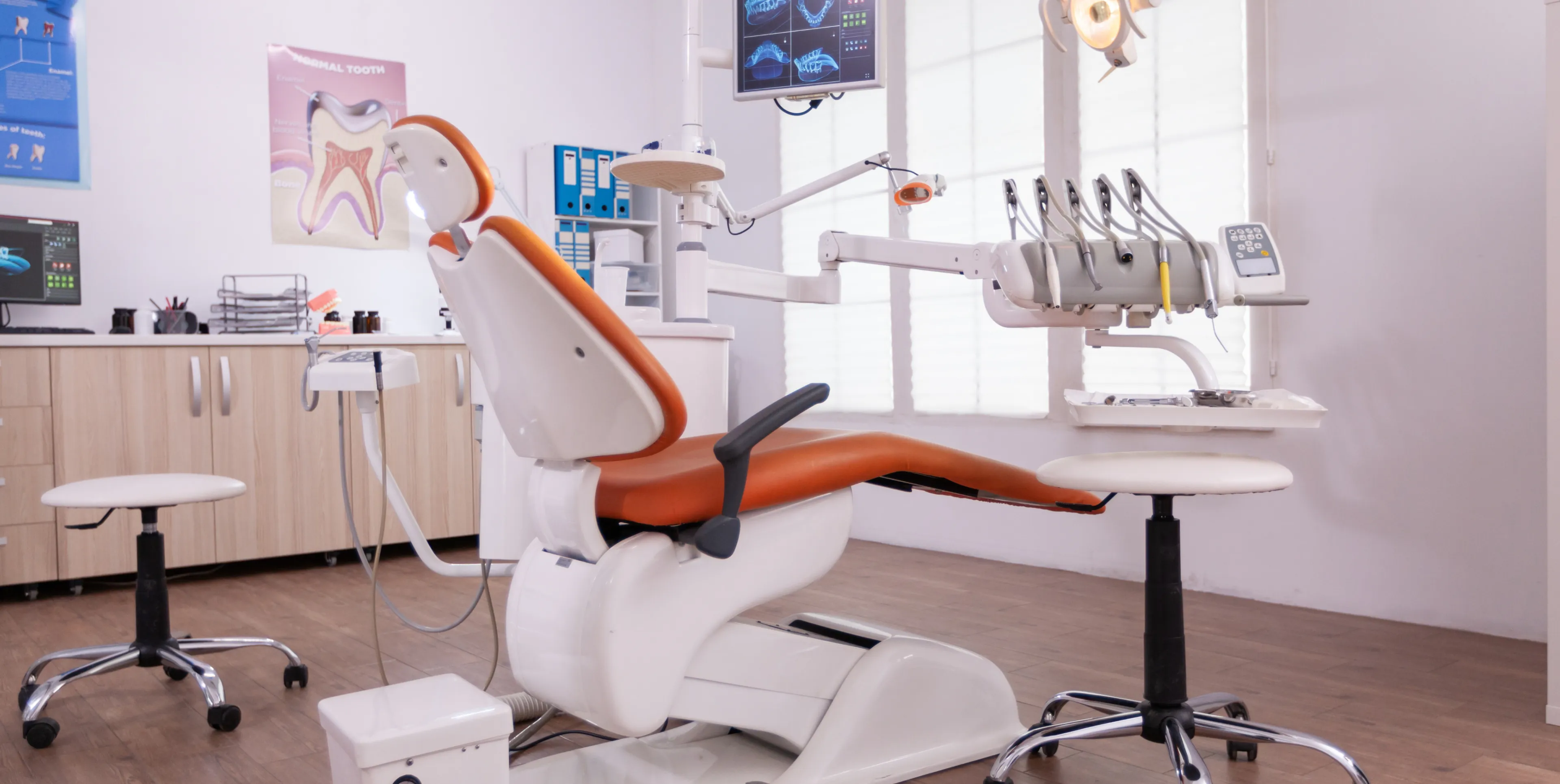 Modern dental examination chair with orange cushioning in a bright clinic room equipped with dental tools and monitors.