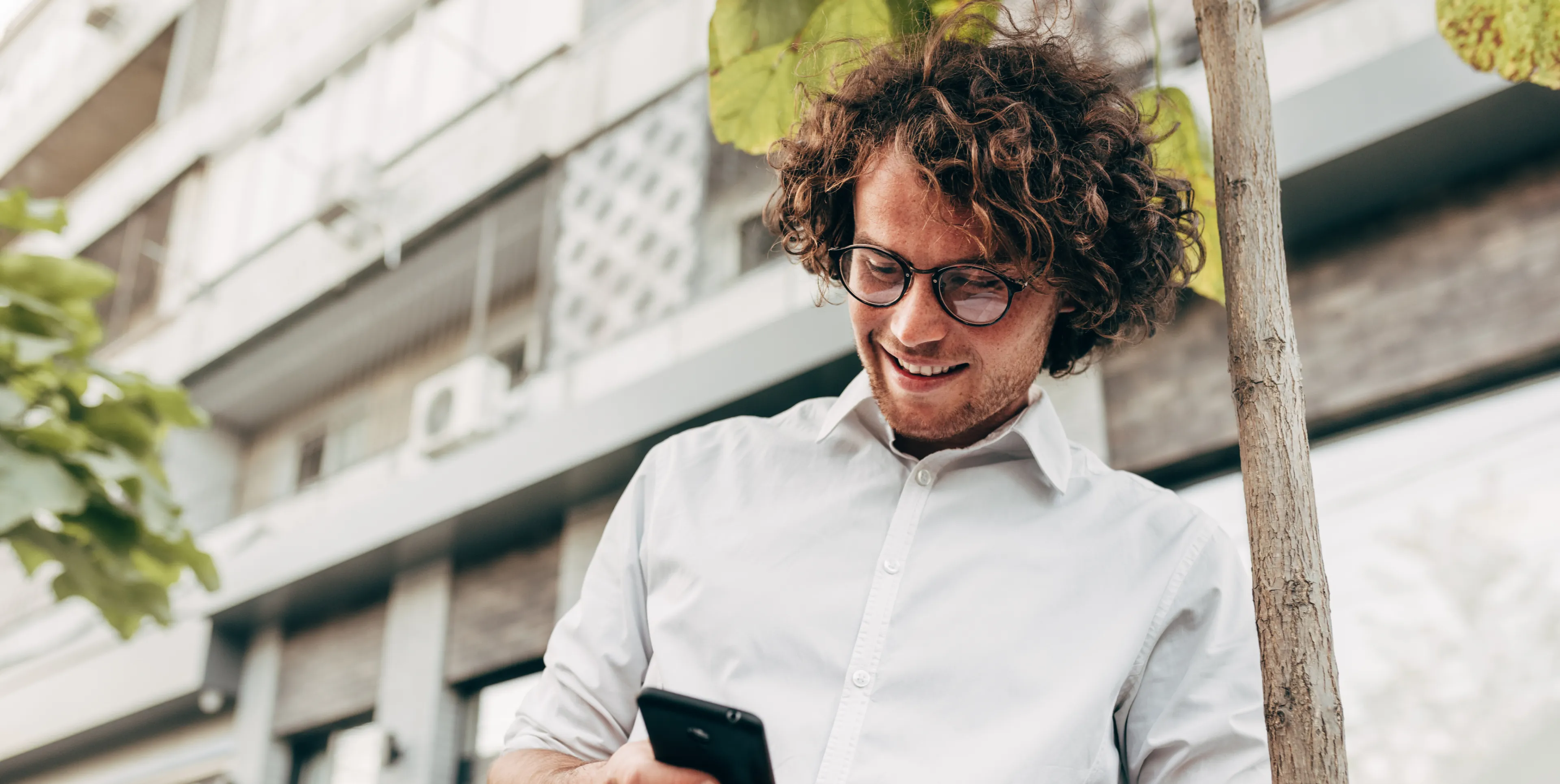 Smiling man with curly hair and glasses looking at smartphone outdoors near a tree and building.