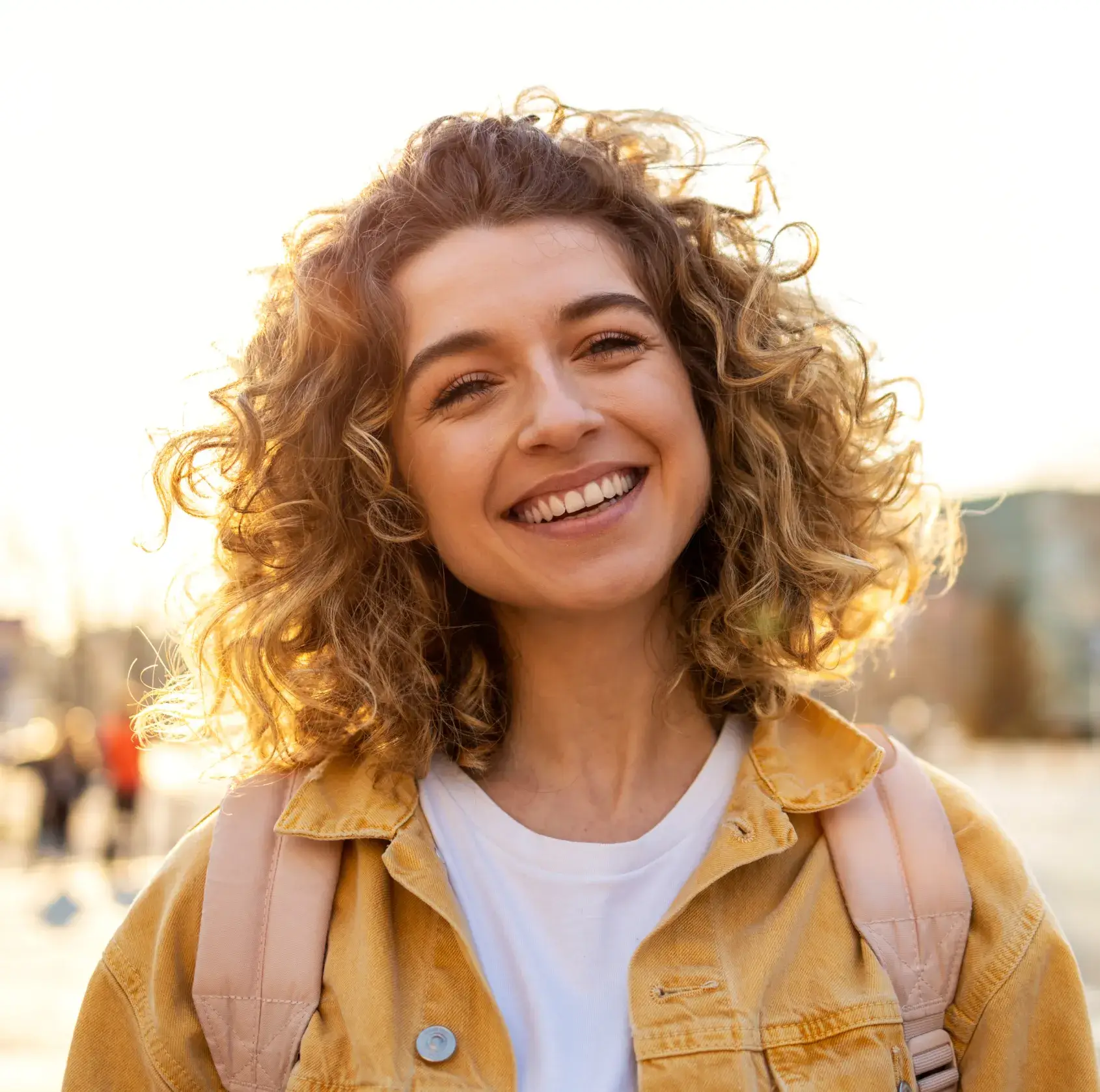 Smiling young woman with curly hair wearing a yellow jacket and backpack outdoors at sunset.