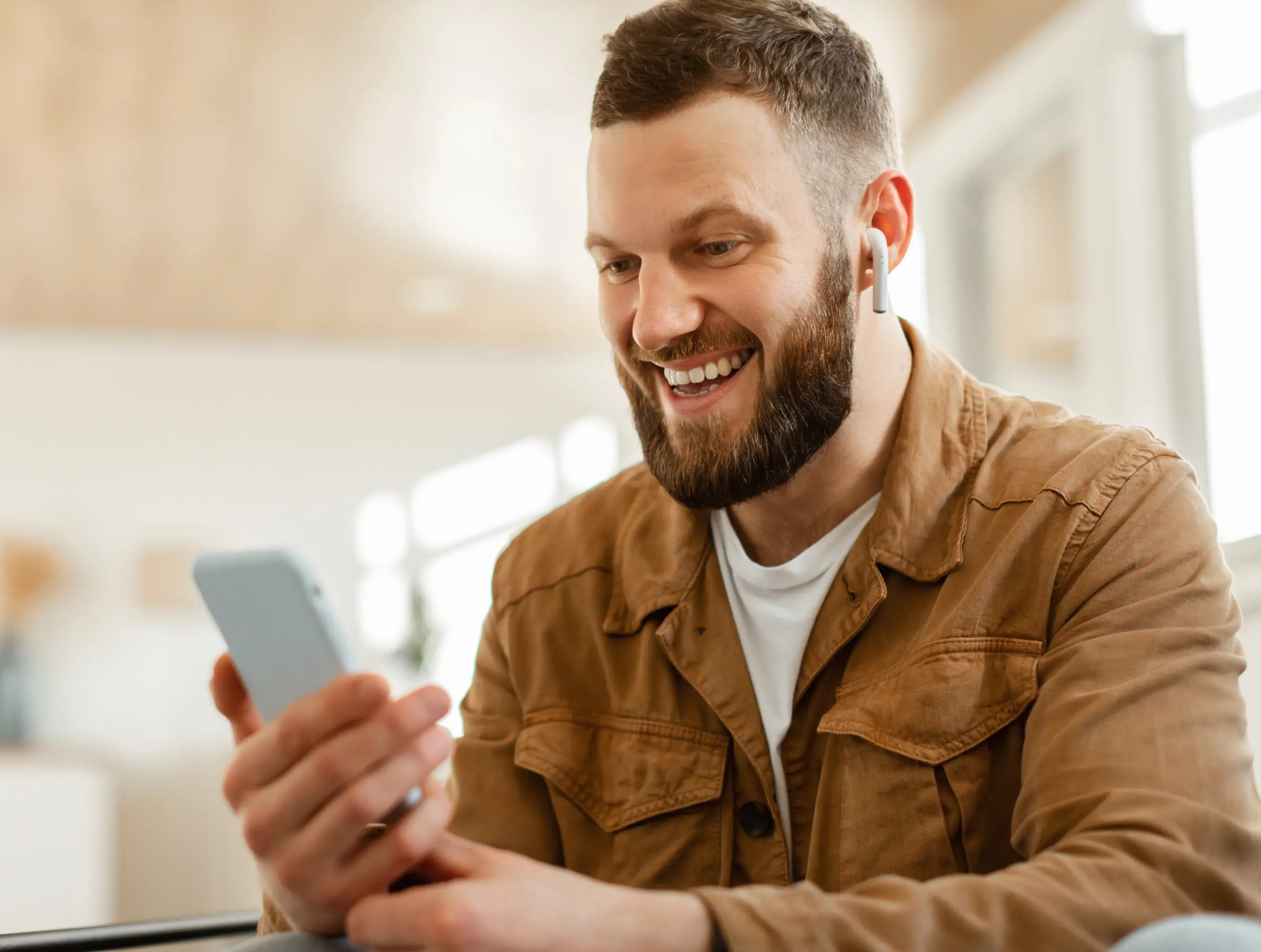 Smiling bearded man wearing wireless earbuds looking at his smartphone inside a bright room.