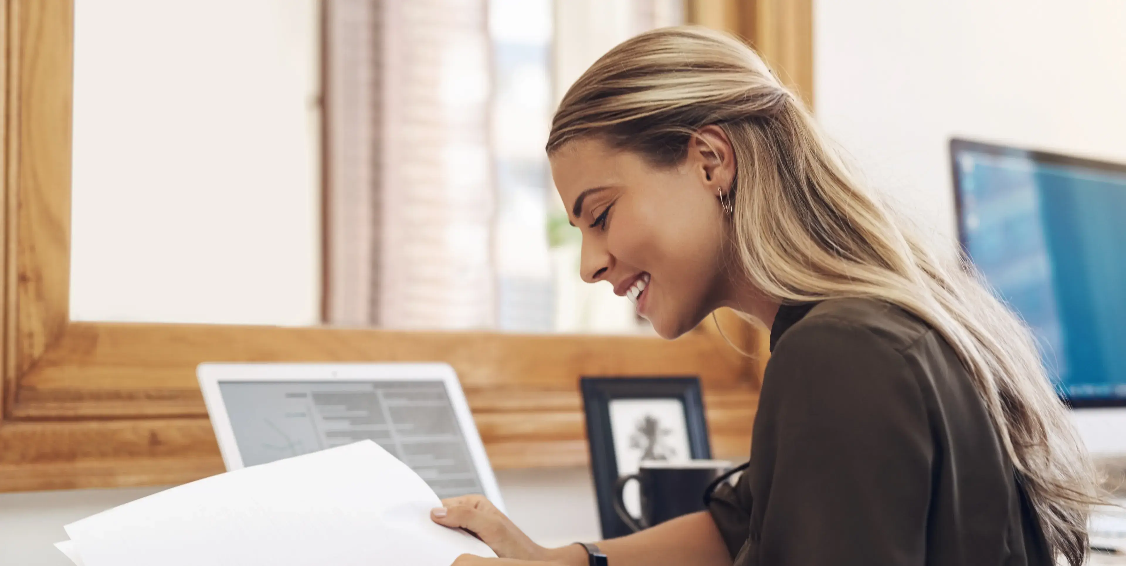 Smiling woman with long blonde hair reviewing papers at a desk with a laptop and computer monitor in the background.