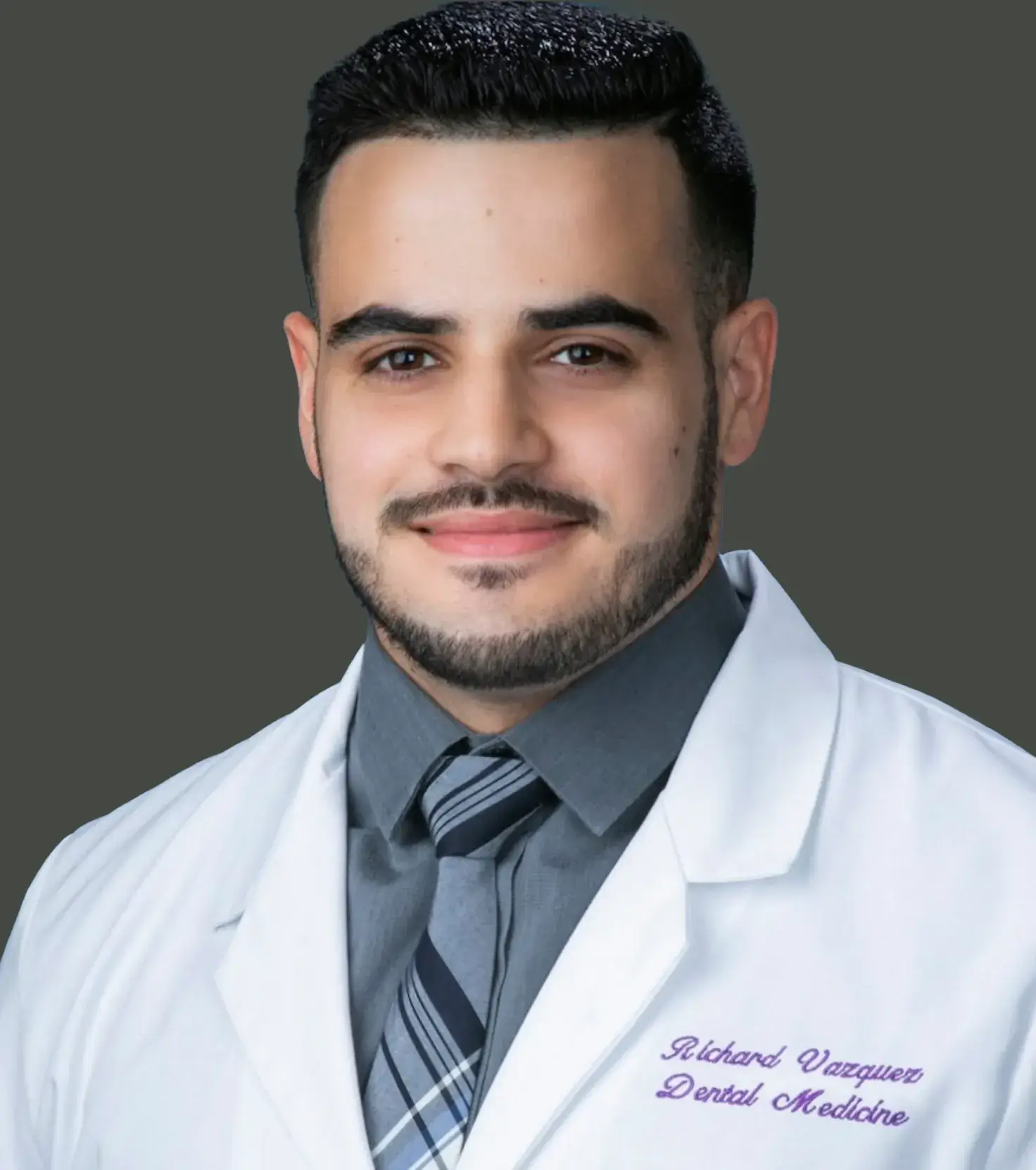 Portrait of a man with dark hair and beard wearing a white lab coat embroidered with the name Richard Vazquez and Dental Medicine, a dark shirt, and a striped tie.