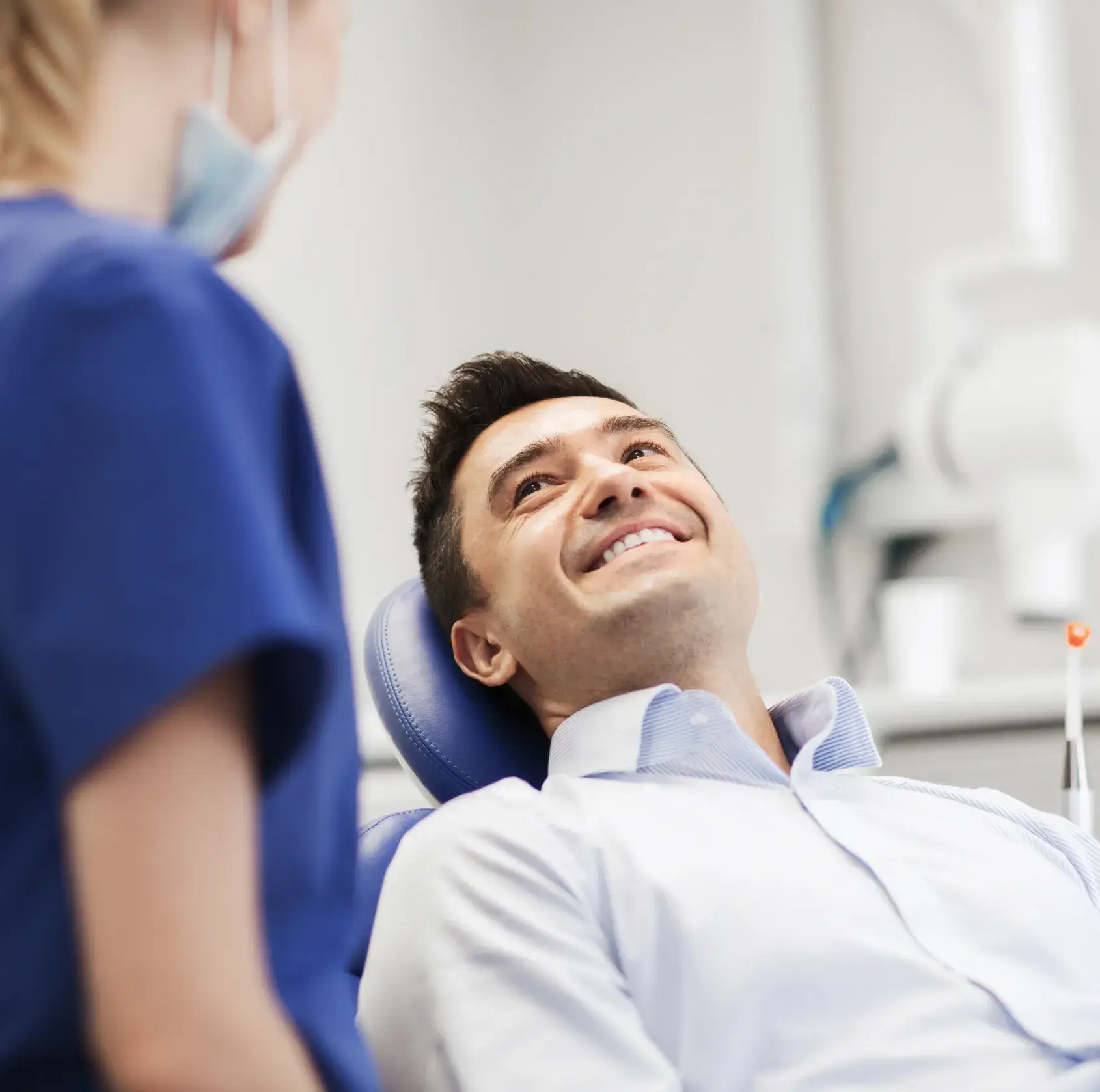 Smiling man reclined in a dental chair looking at a masked dental professional in blue scrubs.