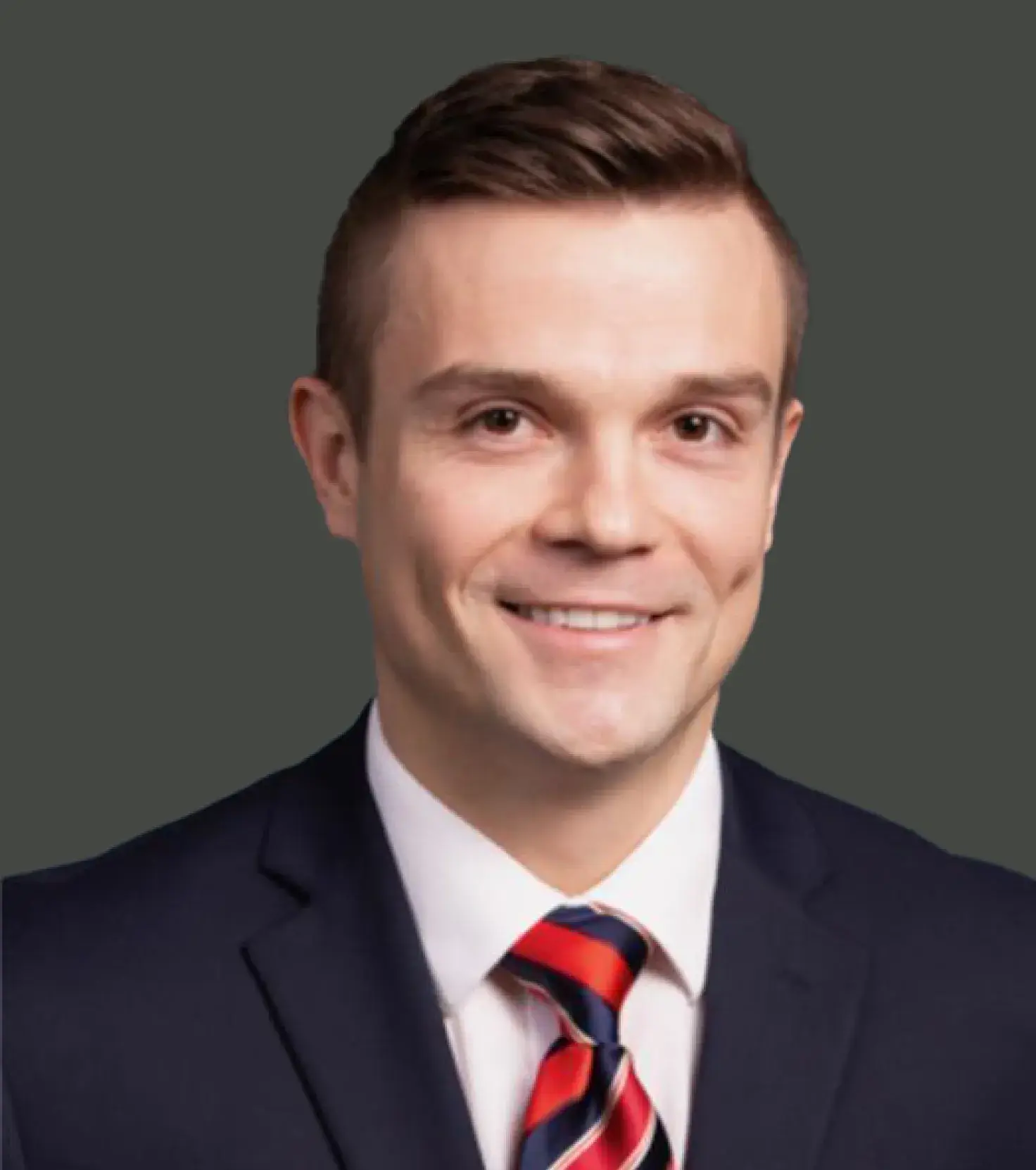 Smiling man in a dark suit with a white shirt and red and blue striped tie against a dark background.