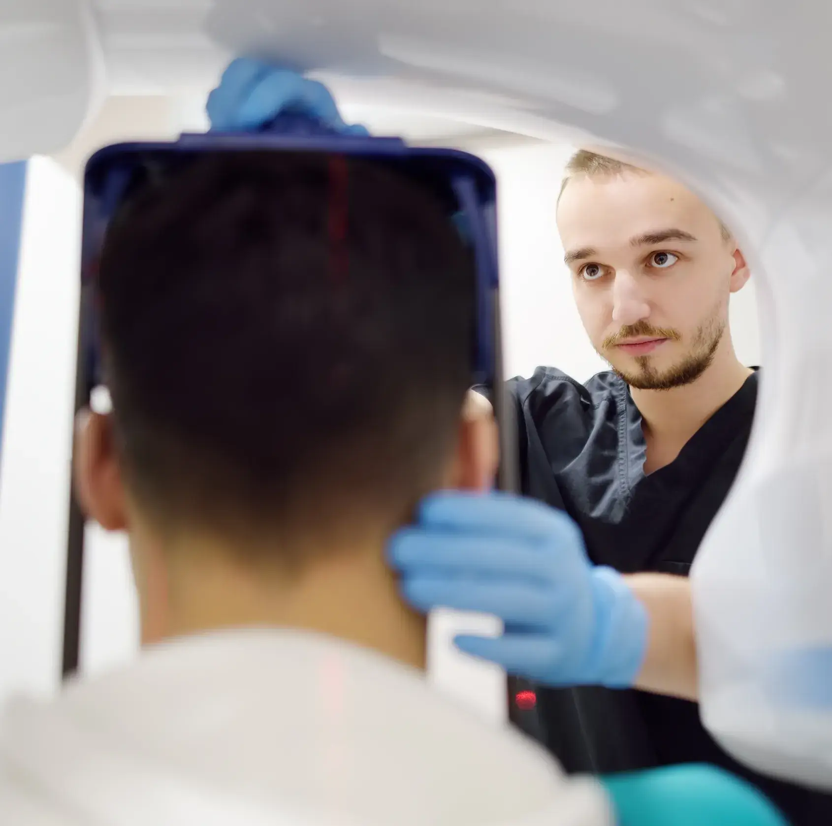 Medical professional wearing blue gloves and black scrubs adjusting equipment for patient during a scan or imaging procedure.