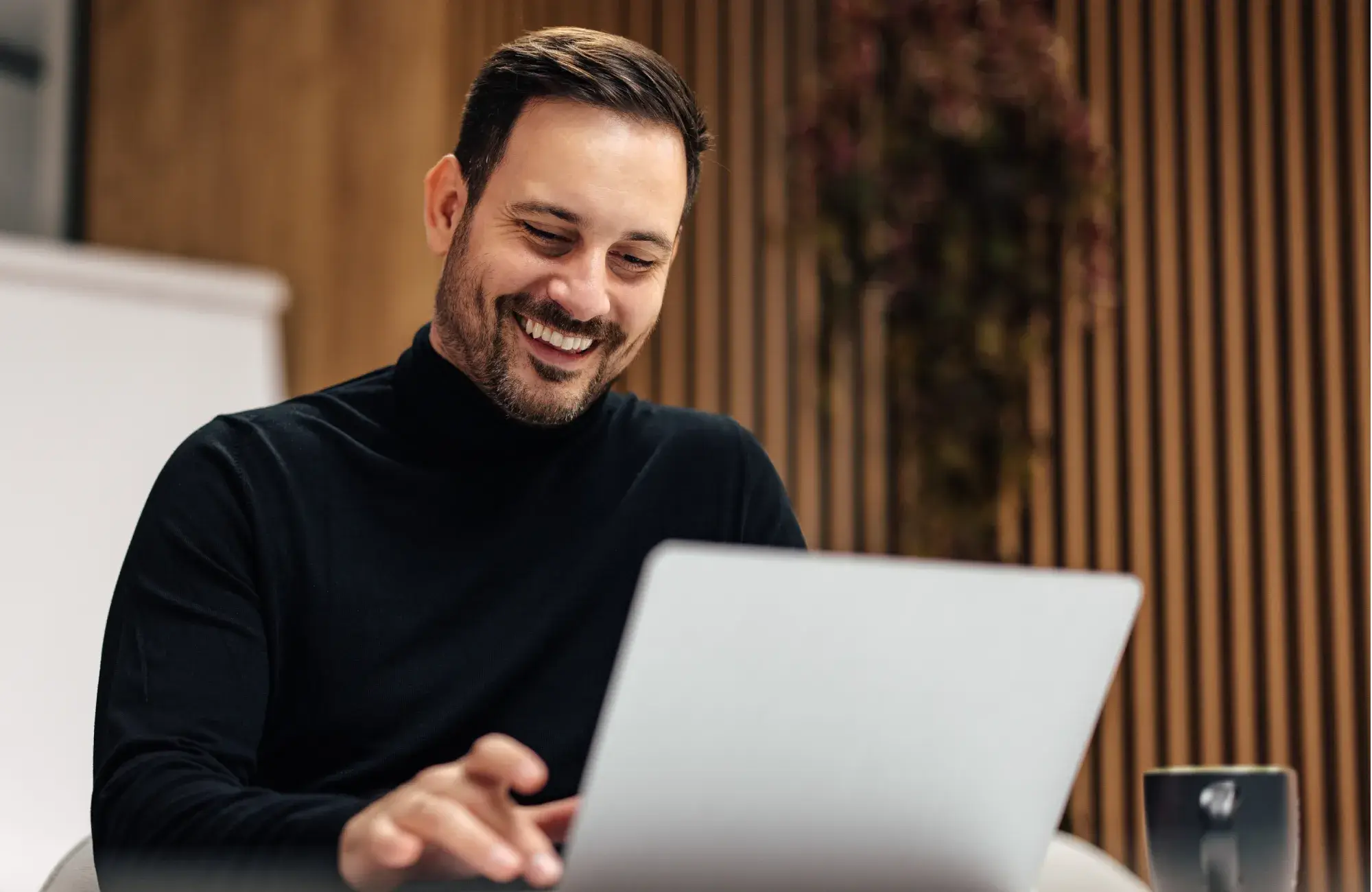 Smiling man in black turtleneck working on a laptop in a modern office setting.