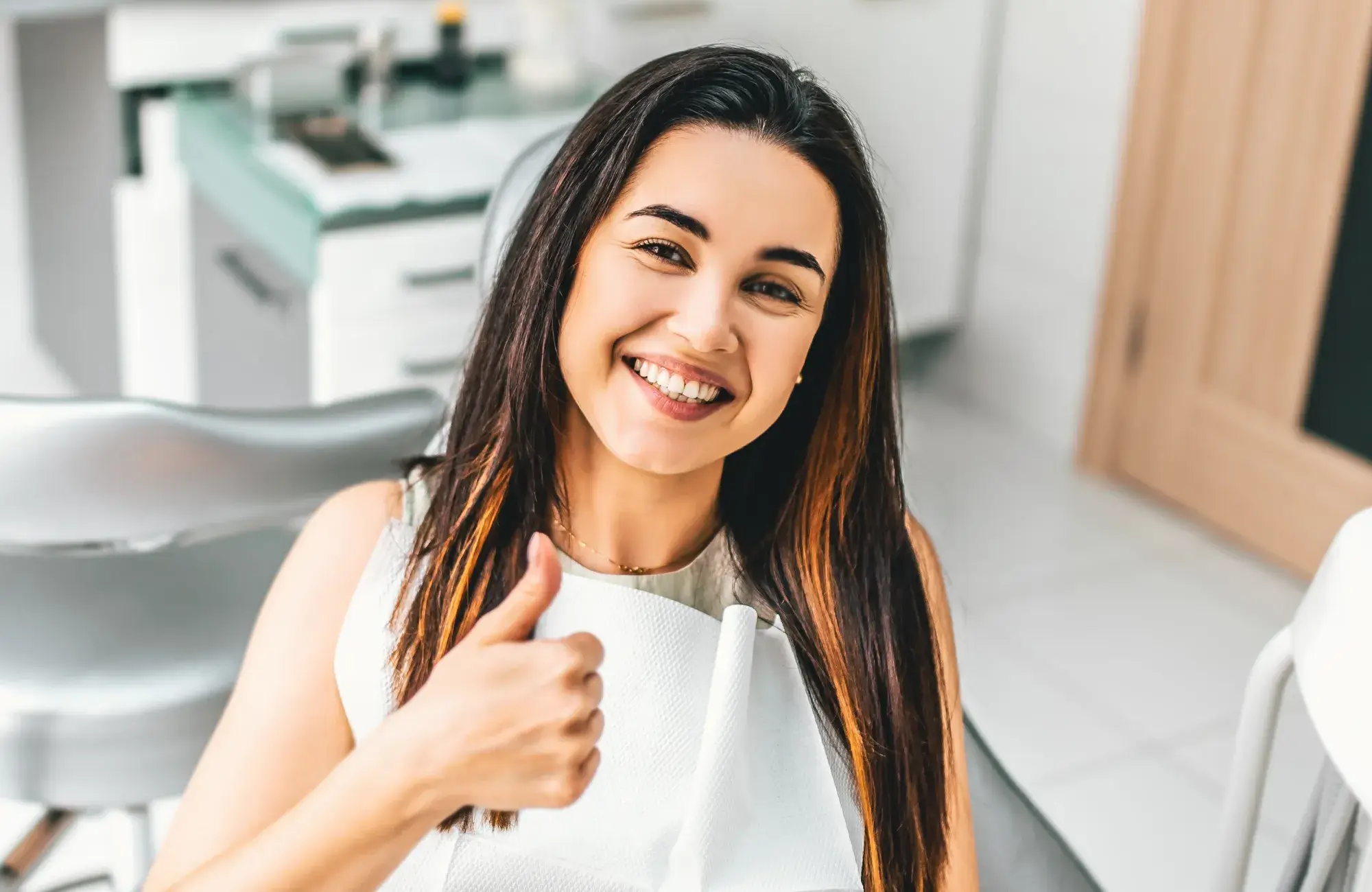 Smiling woman with long dark hair giving a thumbs-up while sitting in a dental chair with a dental bib.