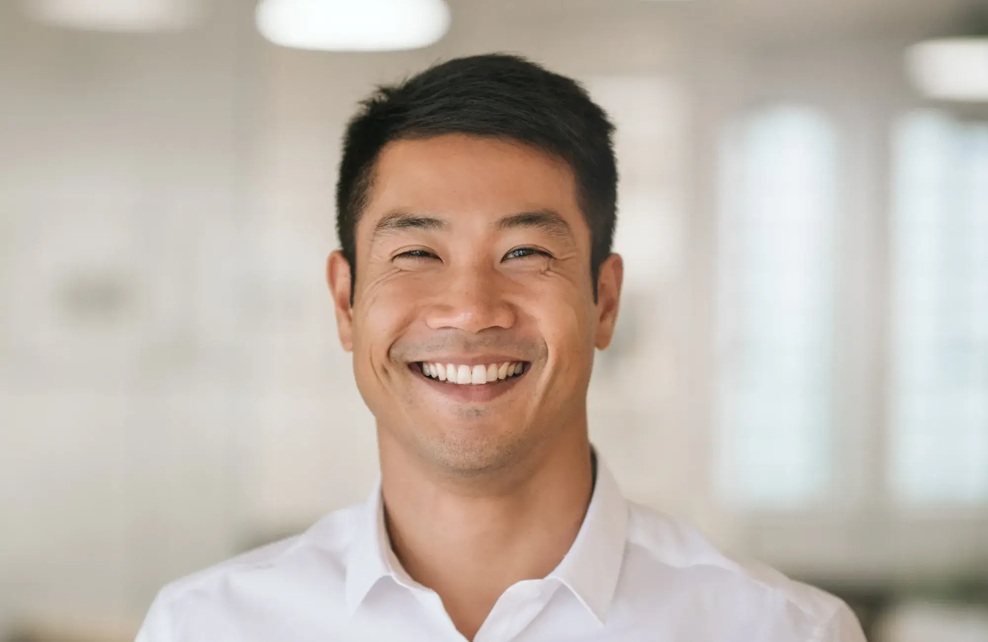 Smiling Asian man wearing a white shirt in a blurred indoor setting.