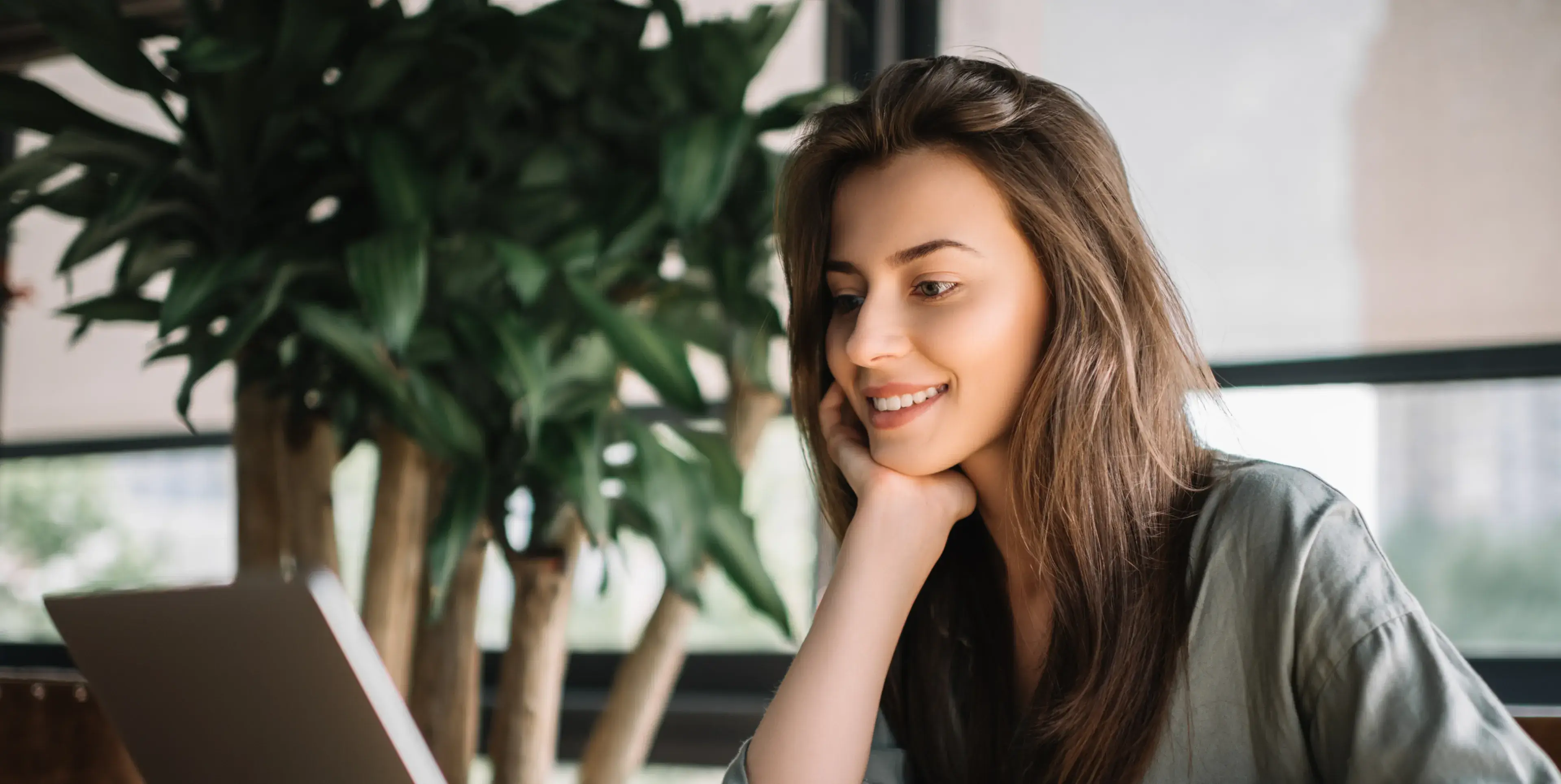 Smiling woman with long hair looking at a laptop in a bright room with large plants in the background.