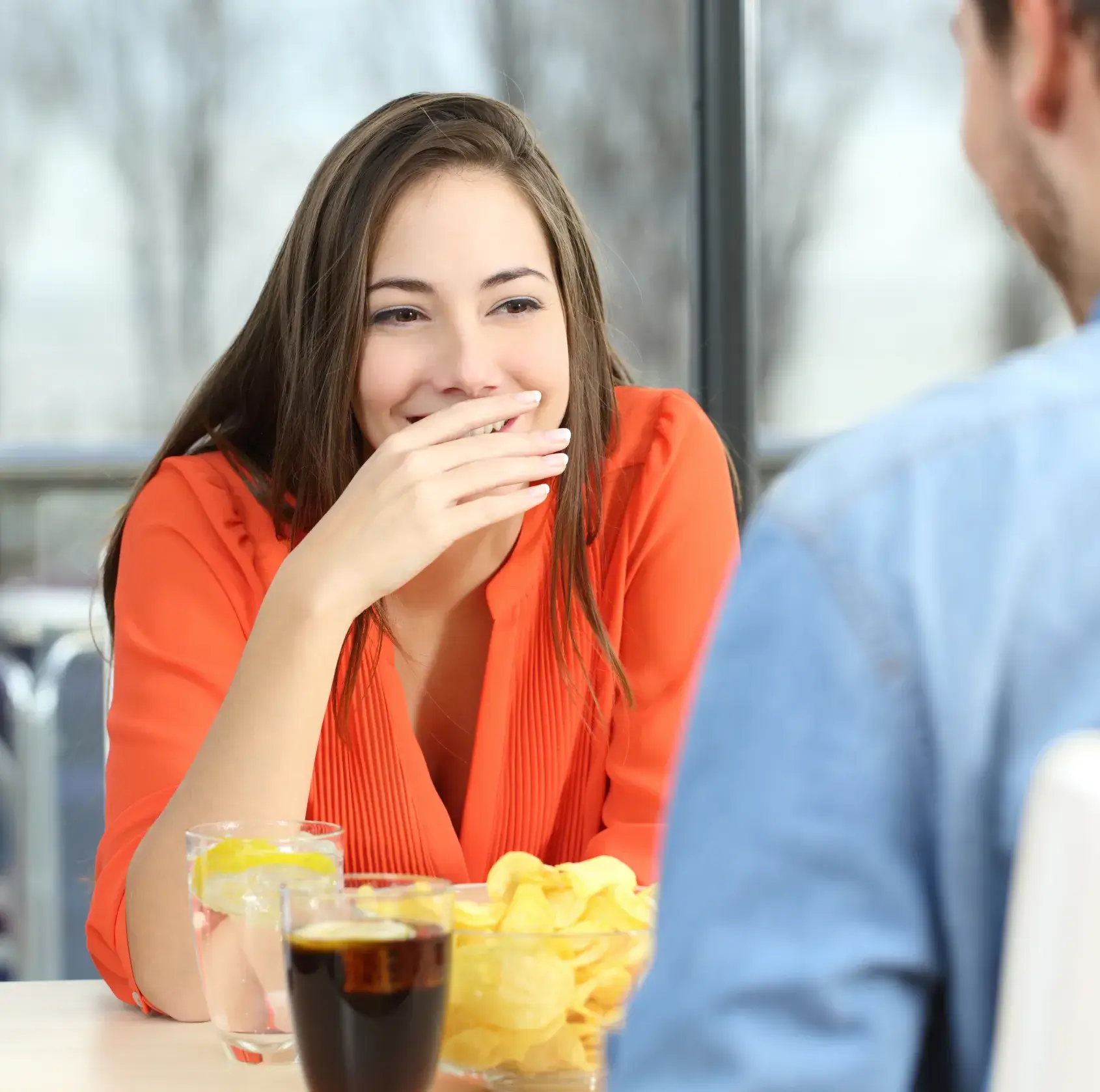 Young woman in an orange blouse laughing with her hand over her mouth while sitting at a table with drinks and potato chips.