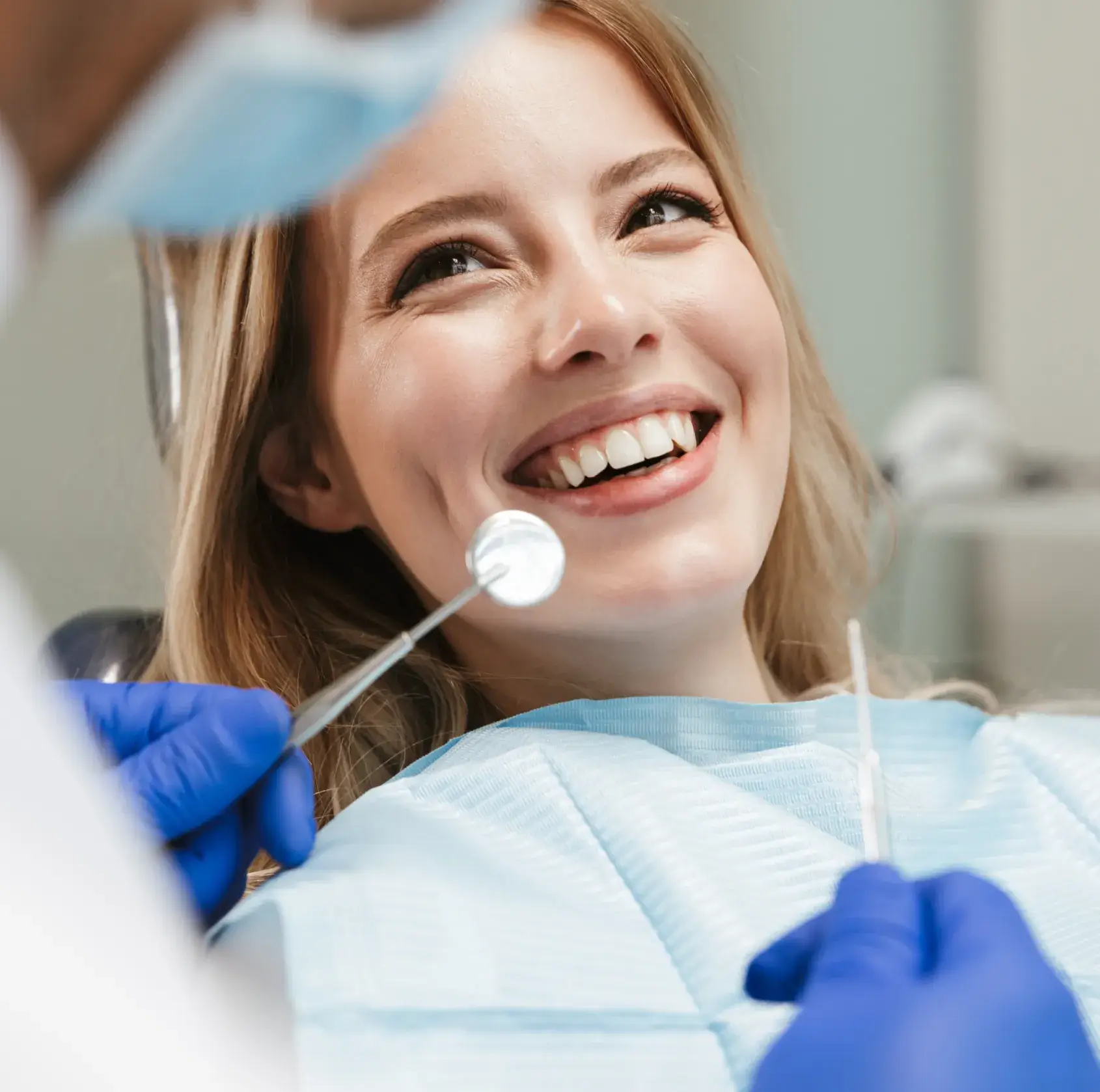 Smiling woman in a dental chair being examined by a dentist wearing blue gloves and holding dental tools.