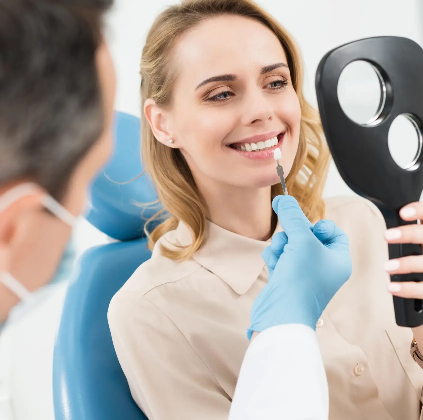 Dentist holding a dental shade guide next to a smiling woman's teeth as she checks her reflection in a black handheld mirror.