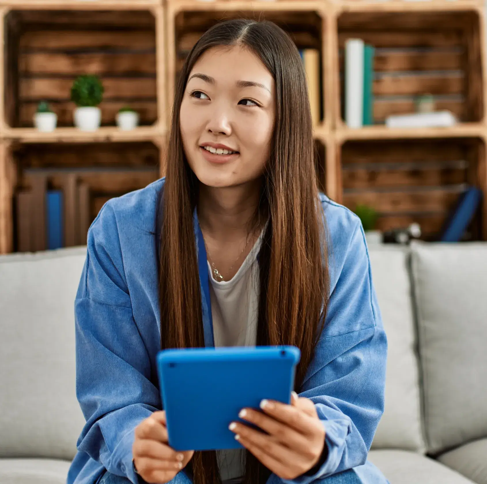 Young woman with long hair in blue jacket holding a blue tablet and looking to the side while sitting on a couch.