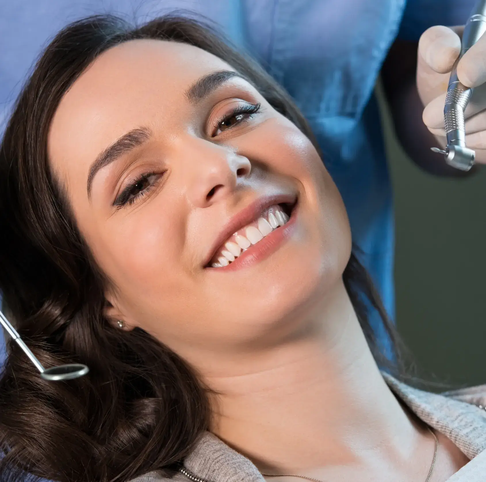 Smiling woman reclined in a dental chair with a dentist holding dental tools nearby.