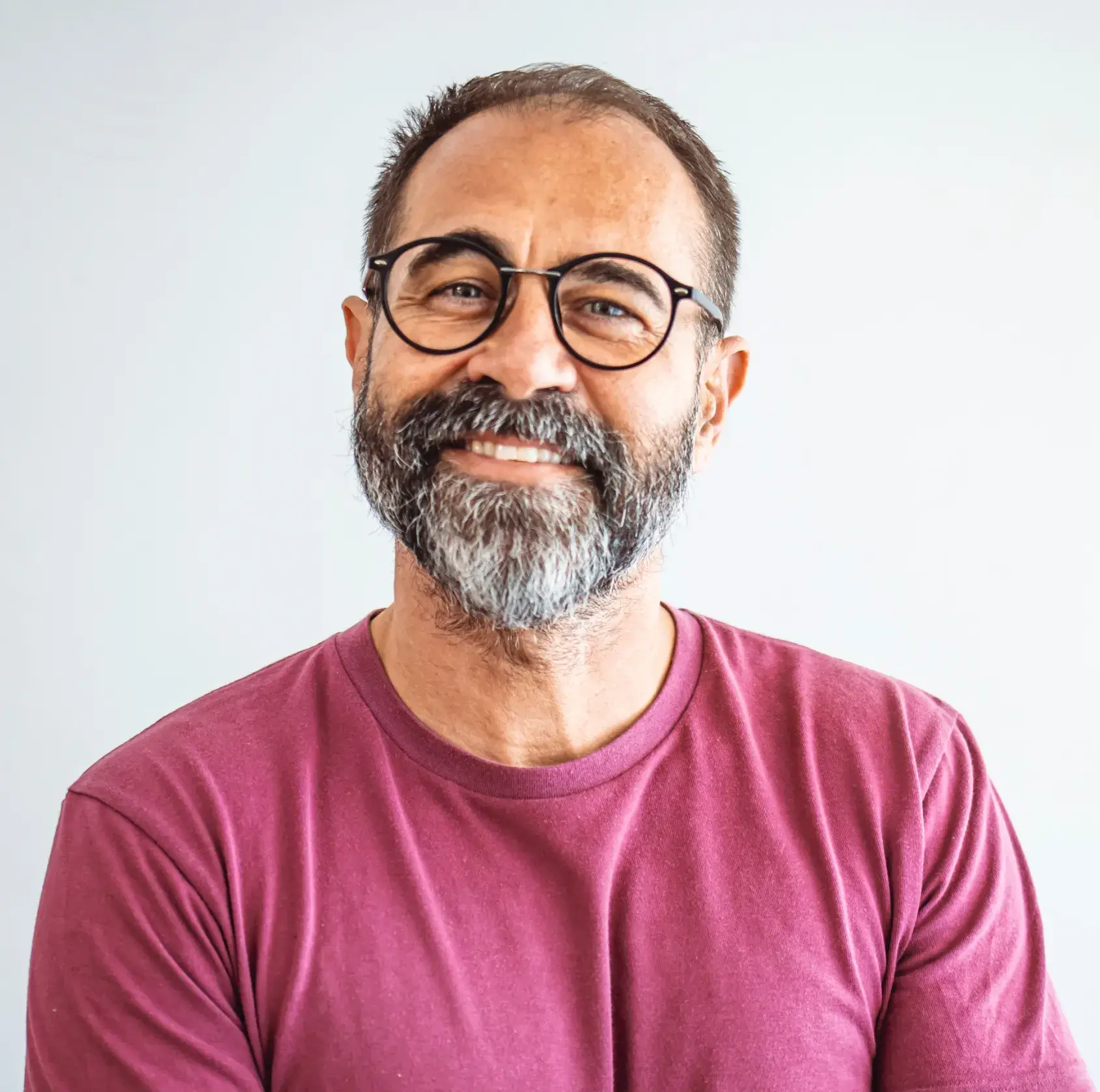 Smiling man with glasses and a gray beard wearing a maroon t-shirt against a light background.