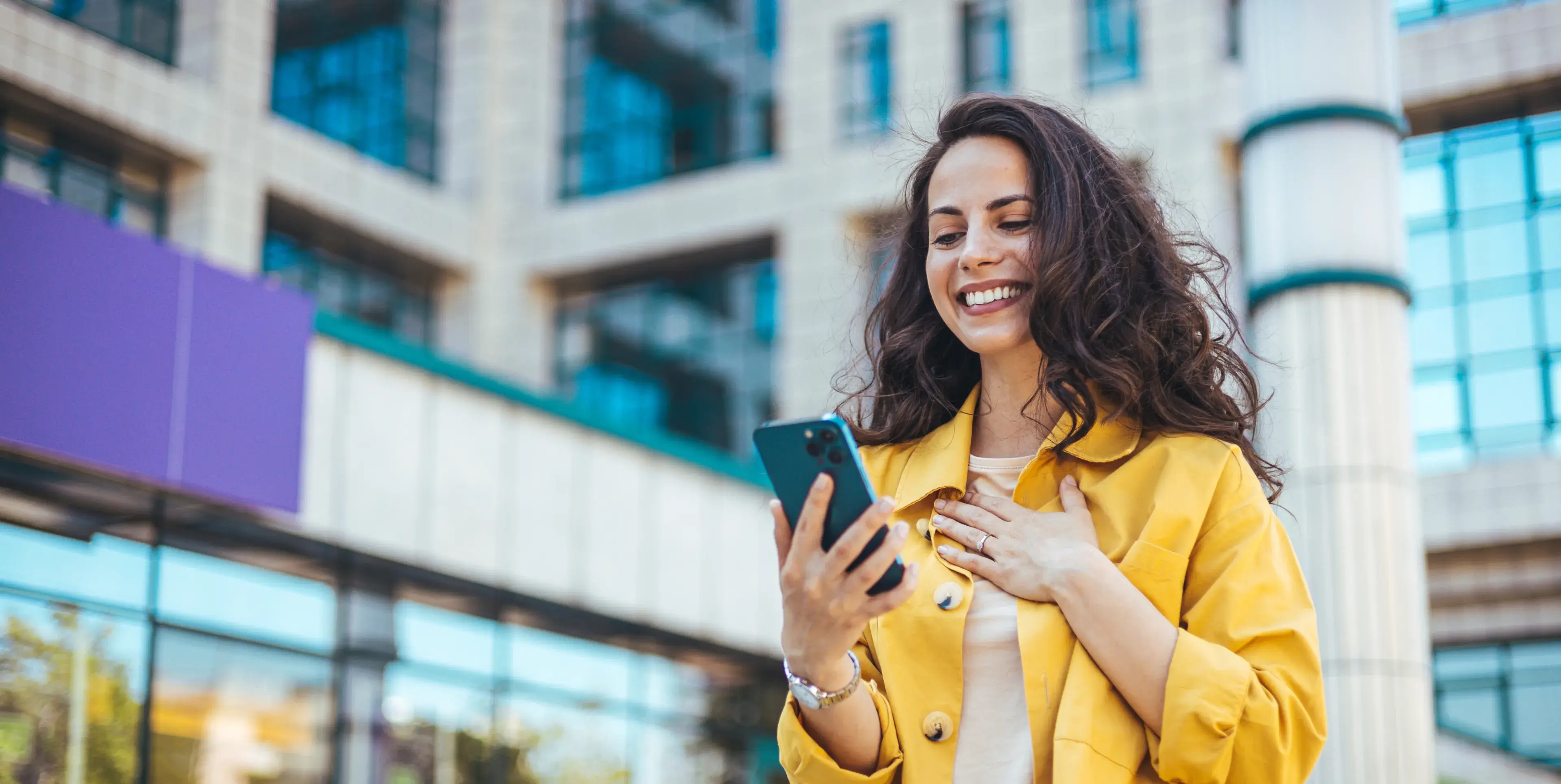 Smiling woman in a yellow jacket looking at her smartphone outdoors in front of a modern glass building.
