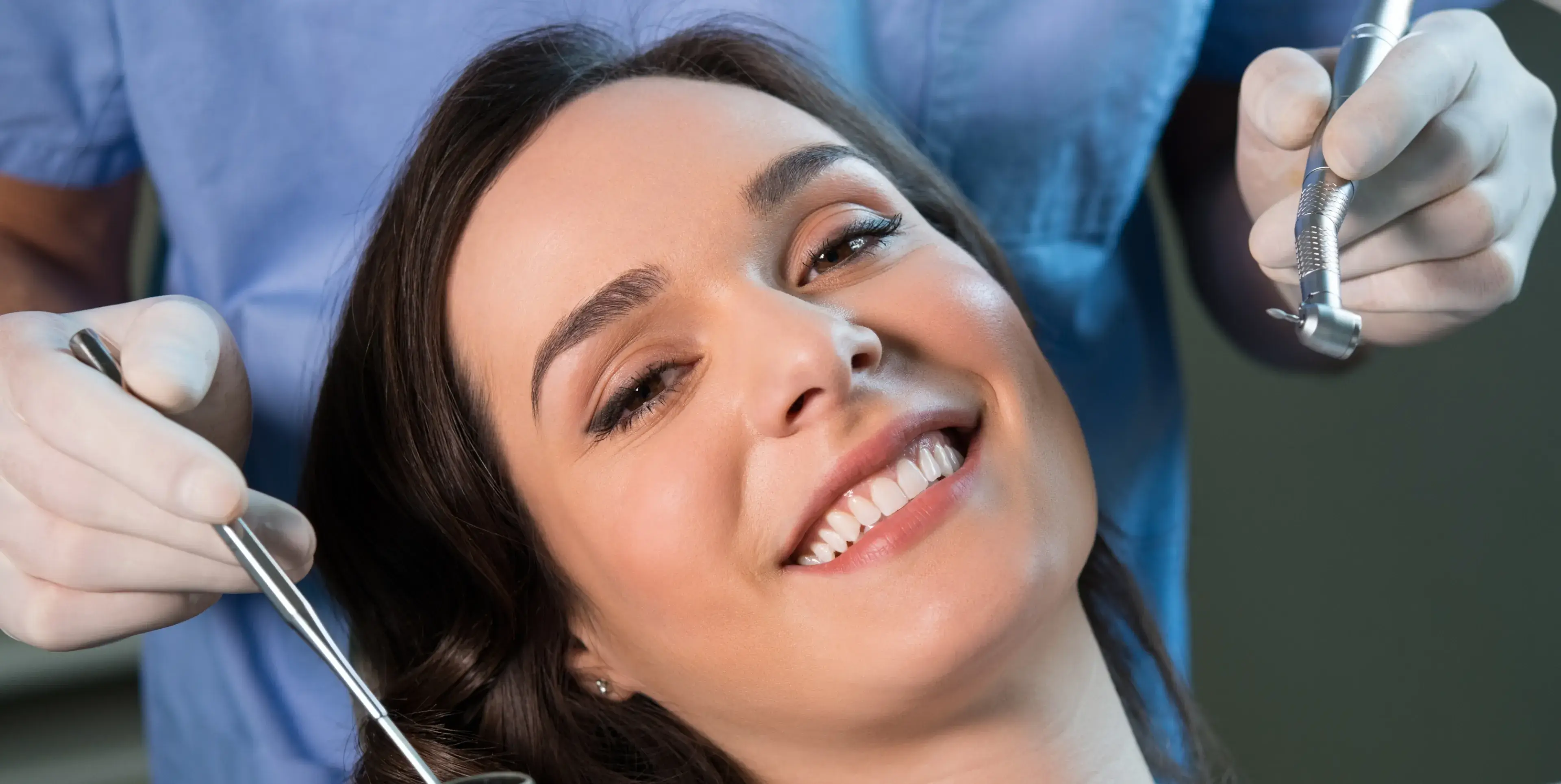 Smiling woman reclined in a dental chair with dentist holding dental tools near her face.