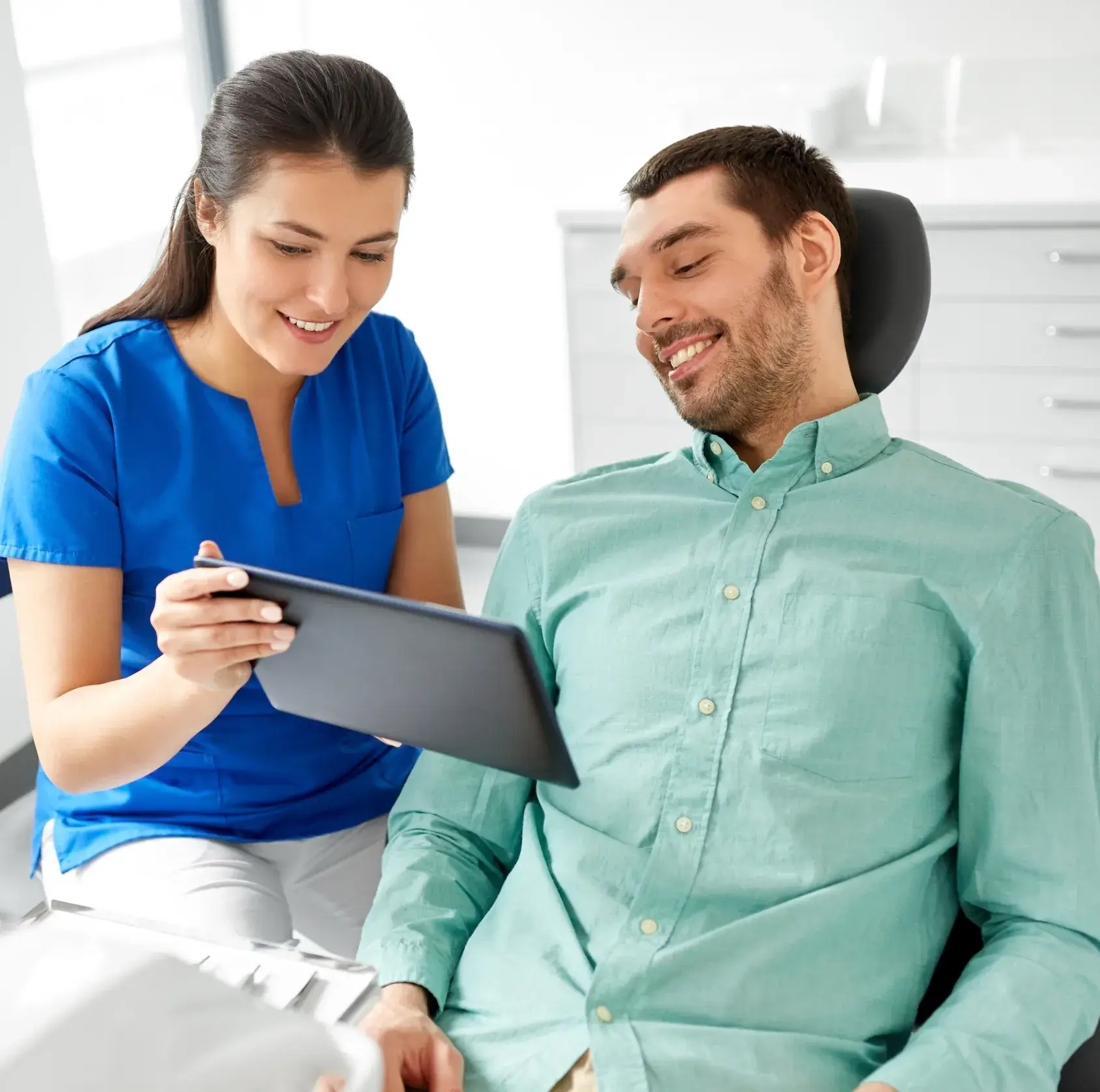 Smiling female healthcare professional showing a tablet to a male patient seated in a dental chair.