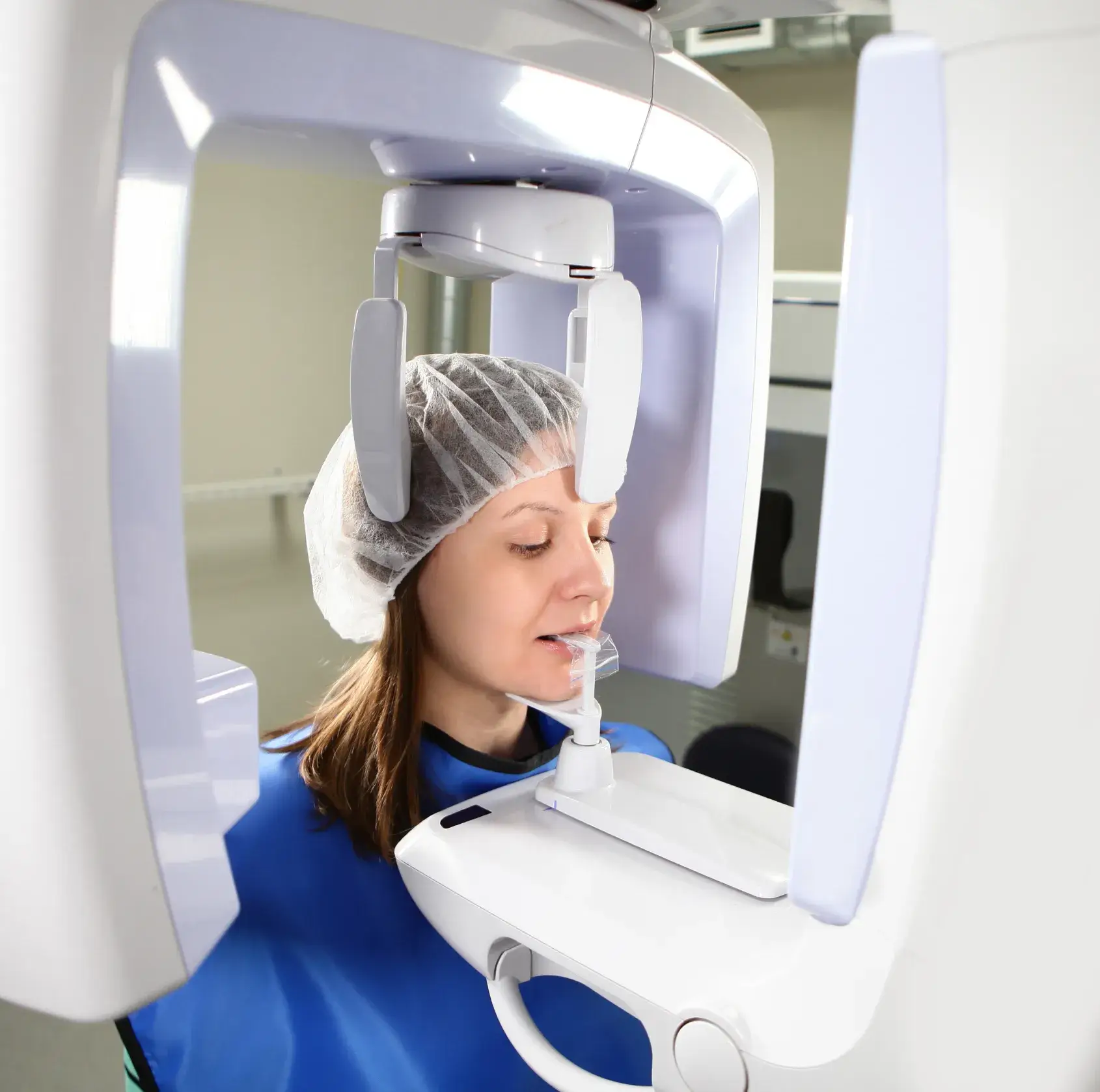 Woman wearing a hairnet and blue protective apron undergoing a dental 3D X-ray scan with a panoramic dental imaging machine.