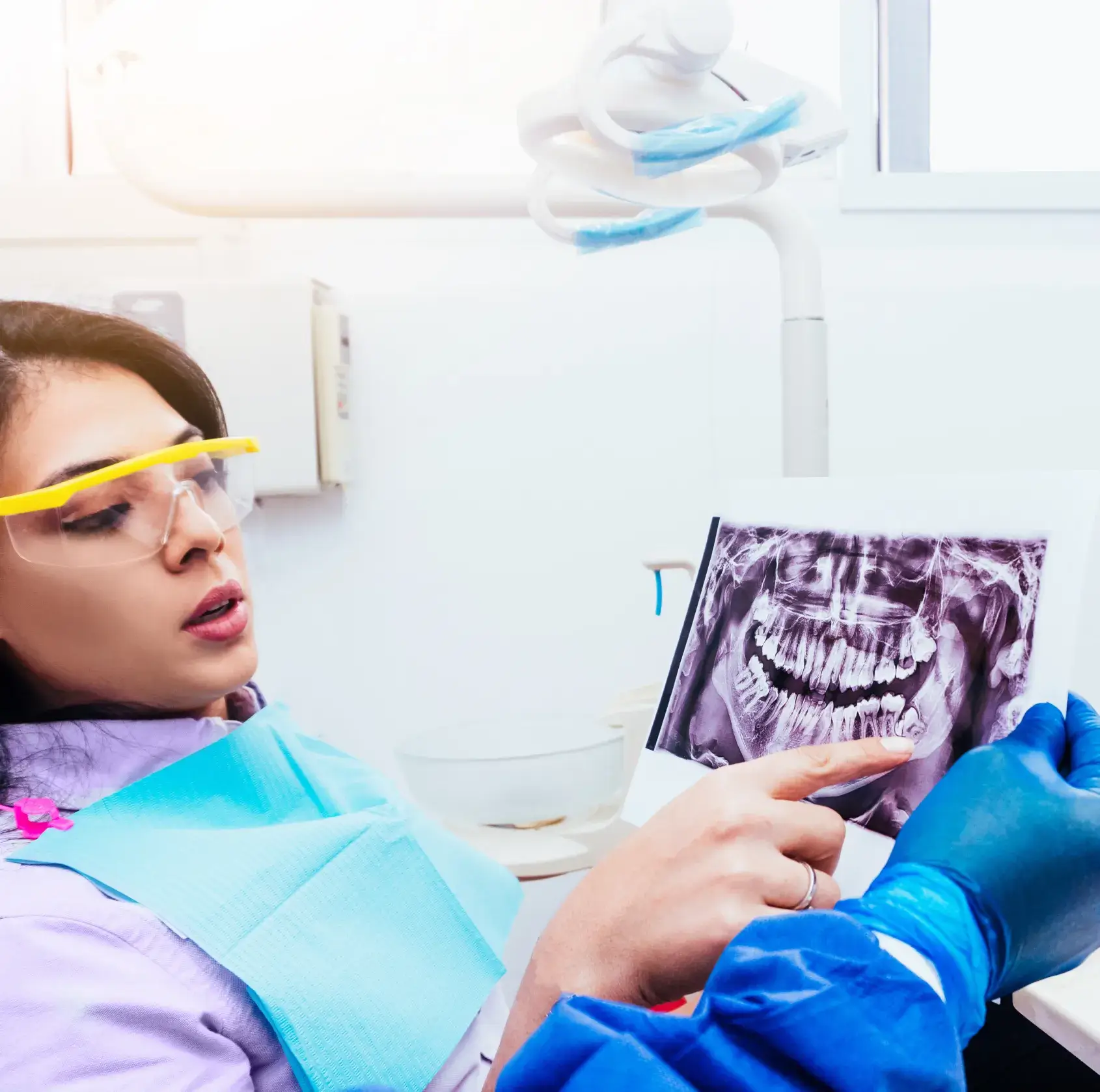 Dentist wearing blue gloves pointing at a dental X-ray while explaining it to a female patient wearing protective glasses and a blue dental bib.