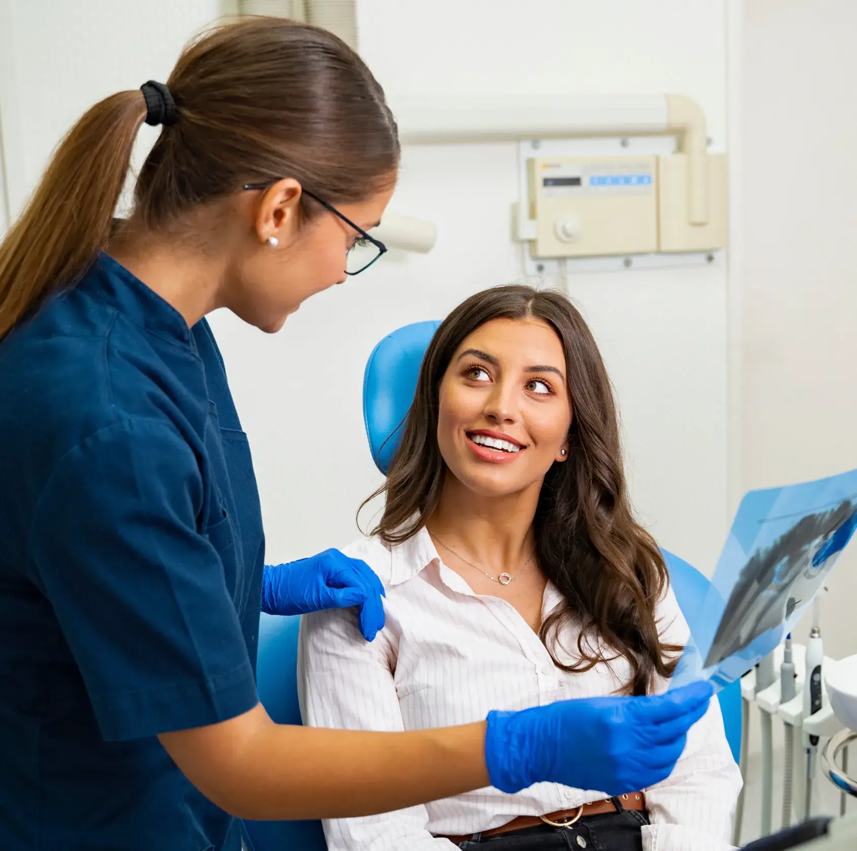 Dentist in blue scrubs showing dental X-rays to a smiling female patient seated in a dental chair.