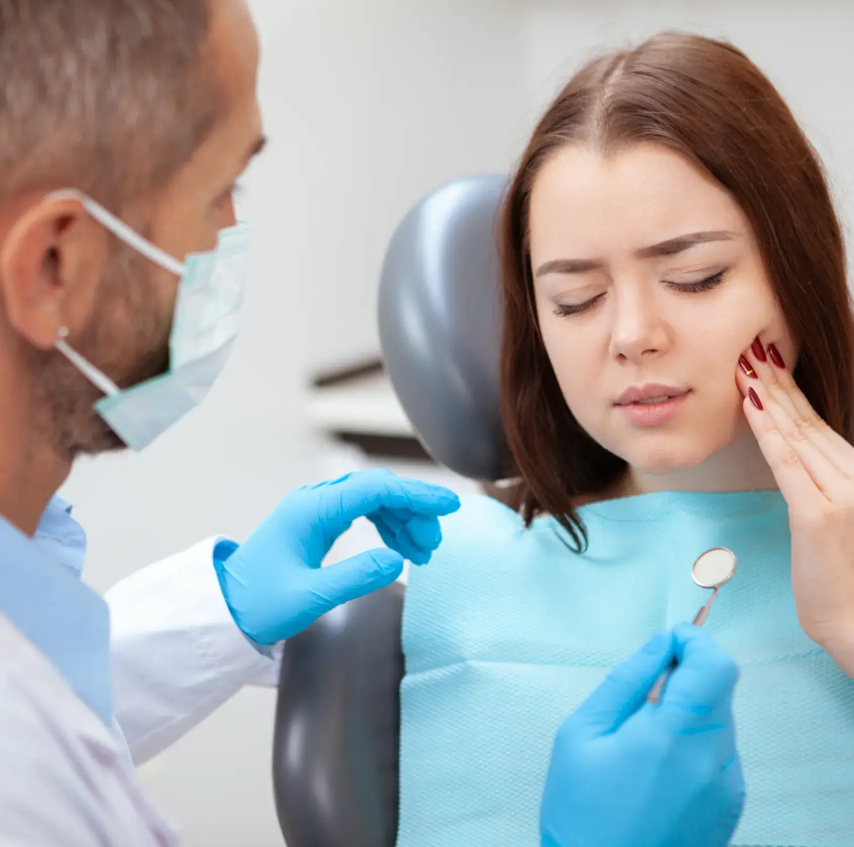 Dentist wearing a mask and blue gloves examining a woman holding her cheek in pain with a dental mirror.