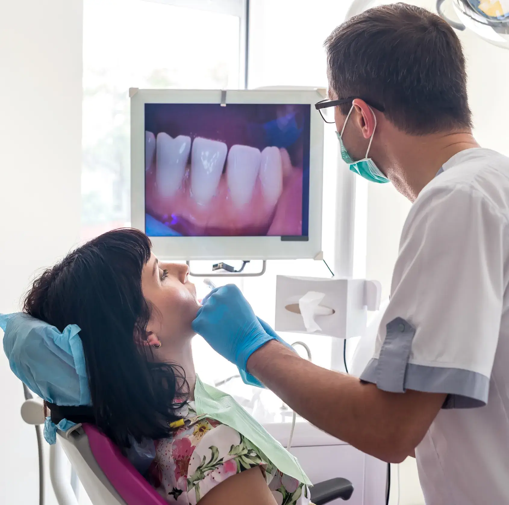 Dentist in mask examining female patient's teeth with dental camera showing close-up of teeth on monitor.