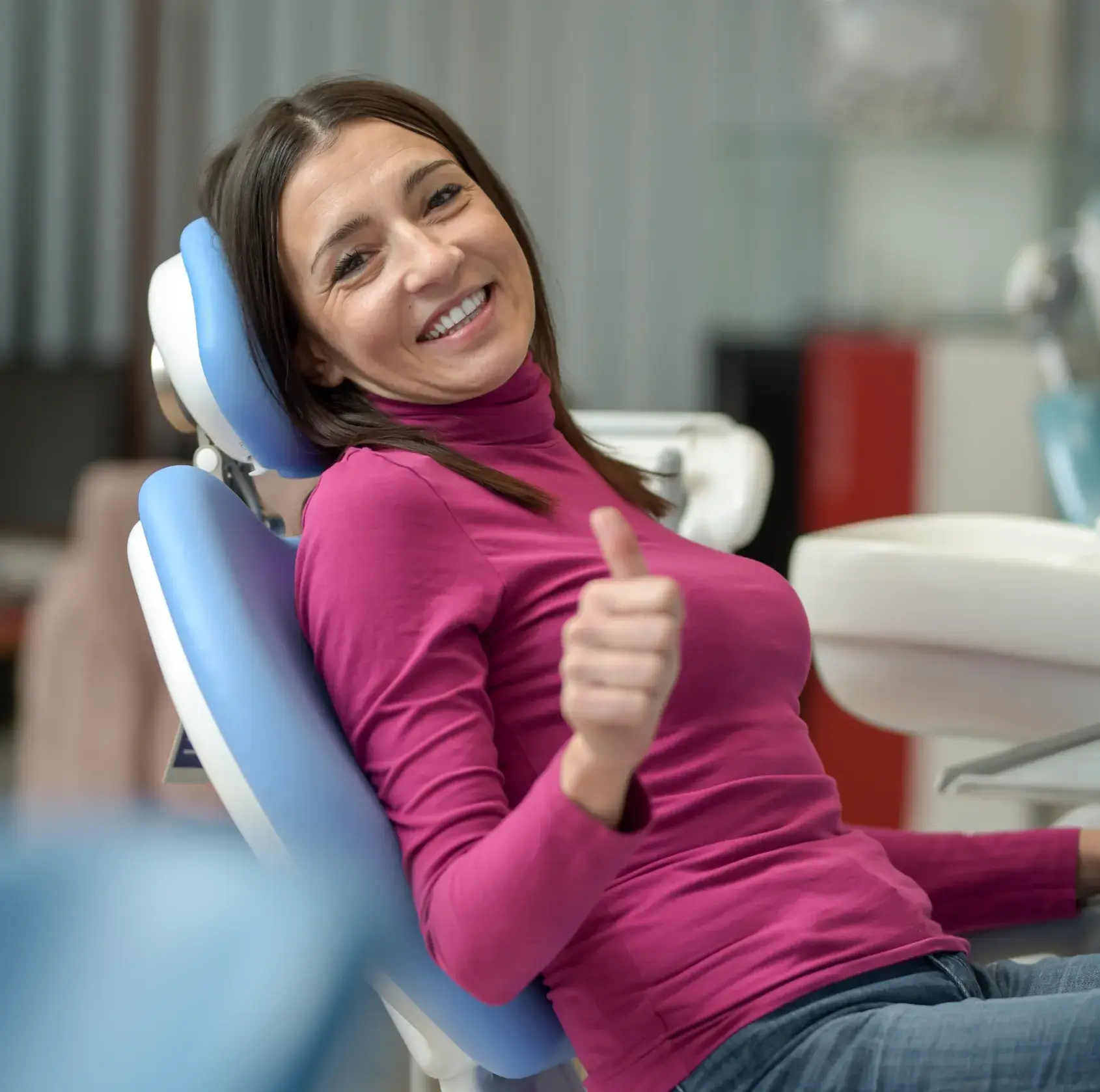 Smiling woman in a pink turtleneck sitting in a dental chair giving a thumbs-up.