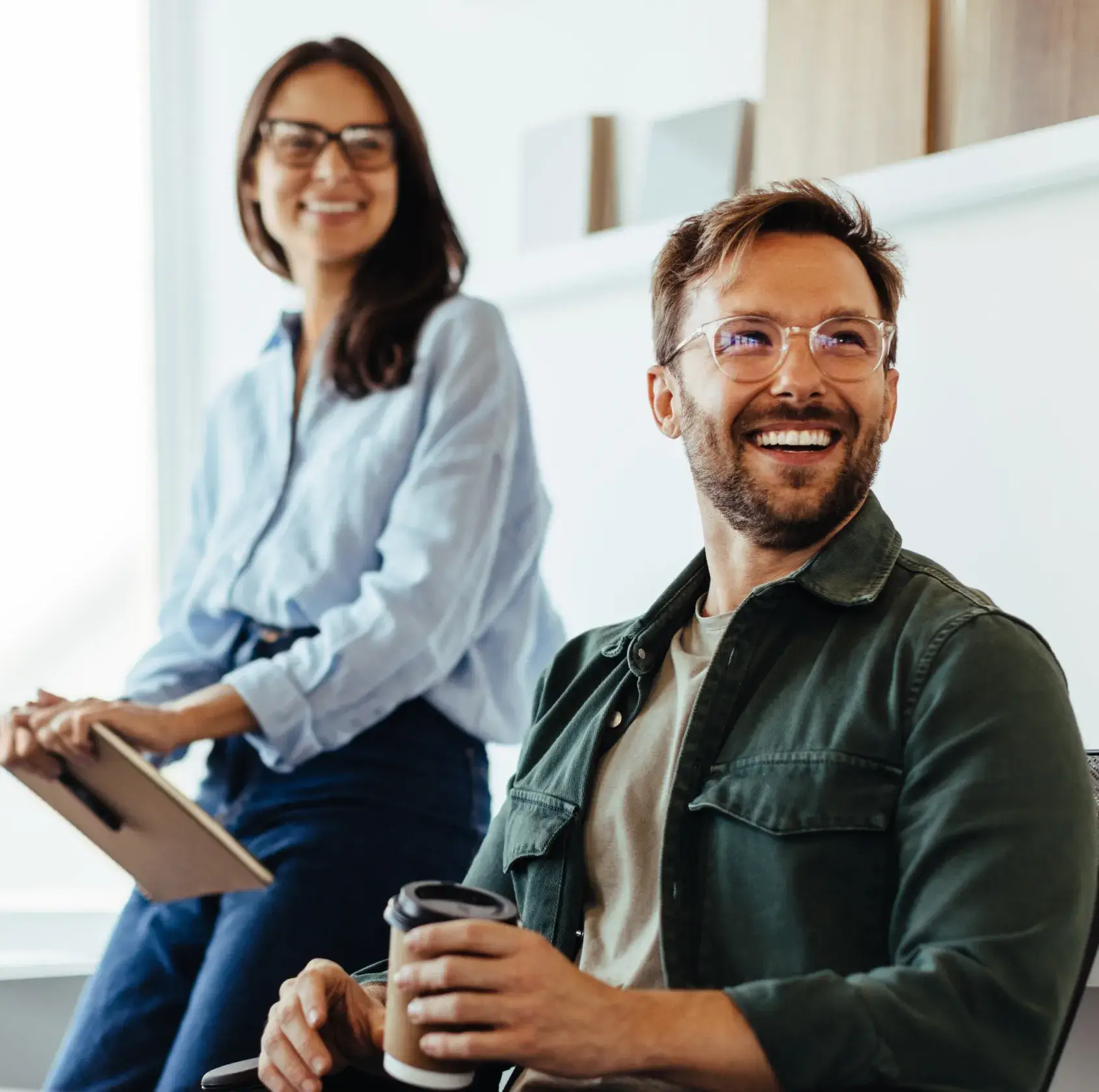 Smiling man holding a coffee cup seated next to a woman with a clipboard in a bright office.