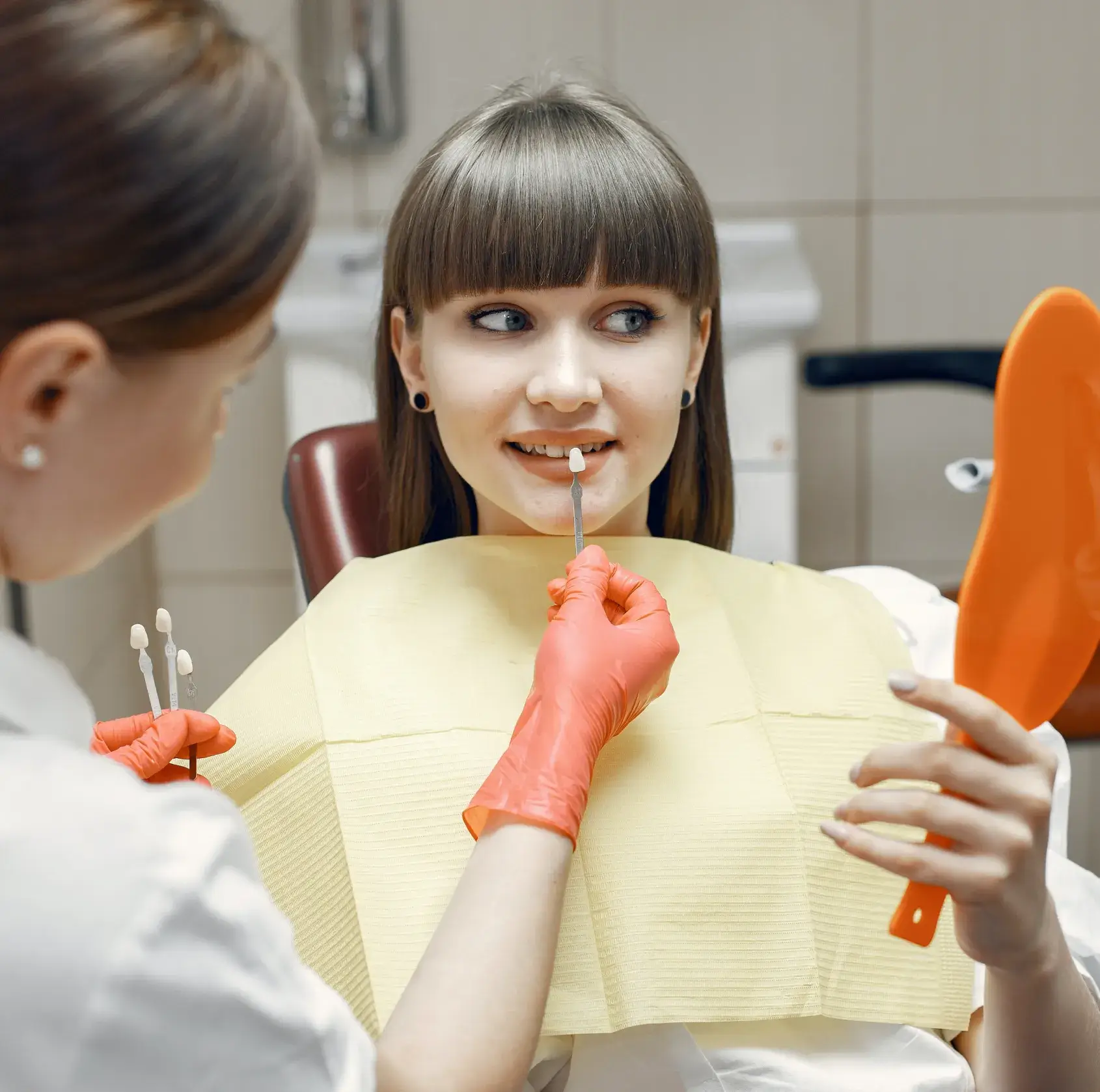 Dentist matching dental veneer shades to a patient's teeth while the patient holds an orange mirror.