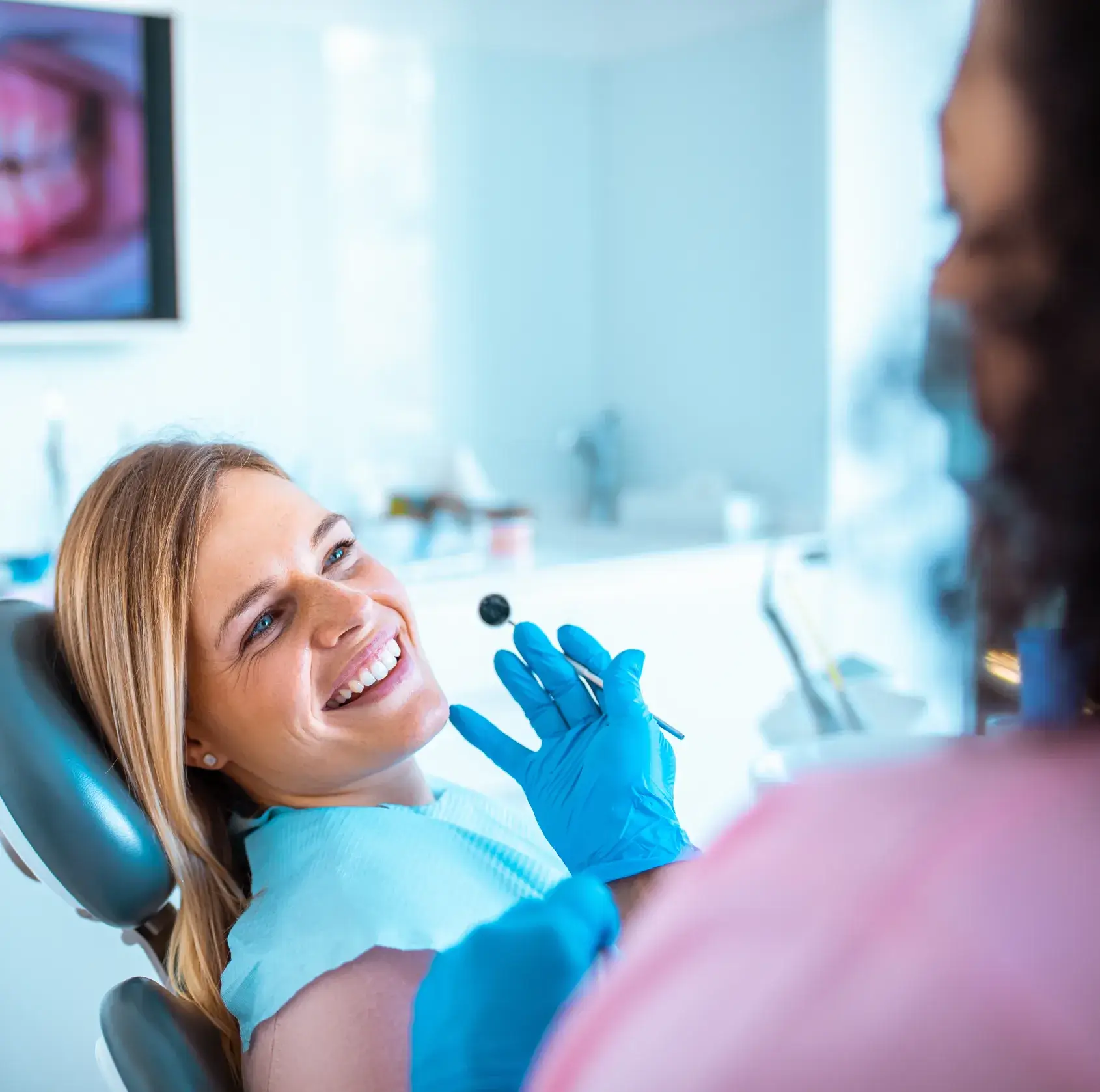 Smiling woman sitting in a dental chair looking at a dentist holding a dental mirror.