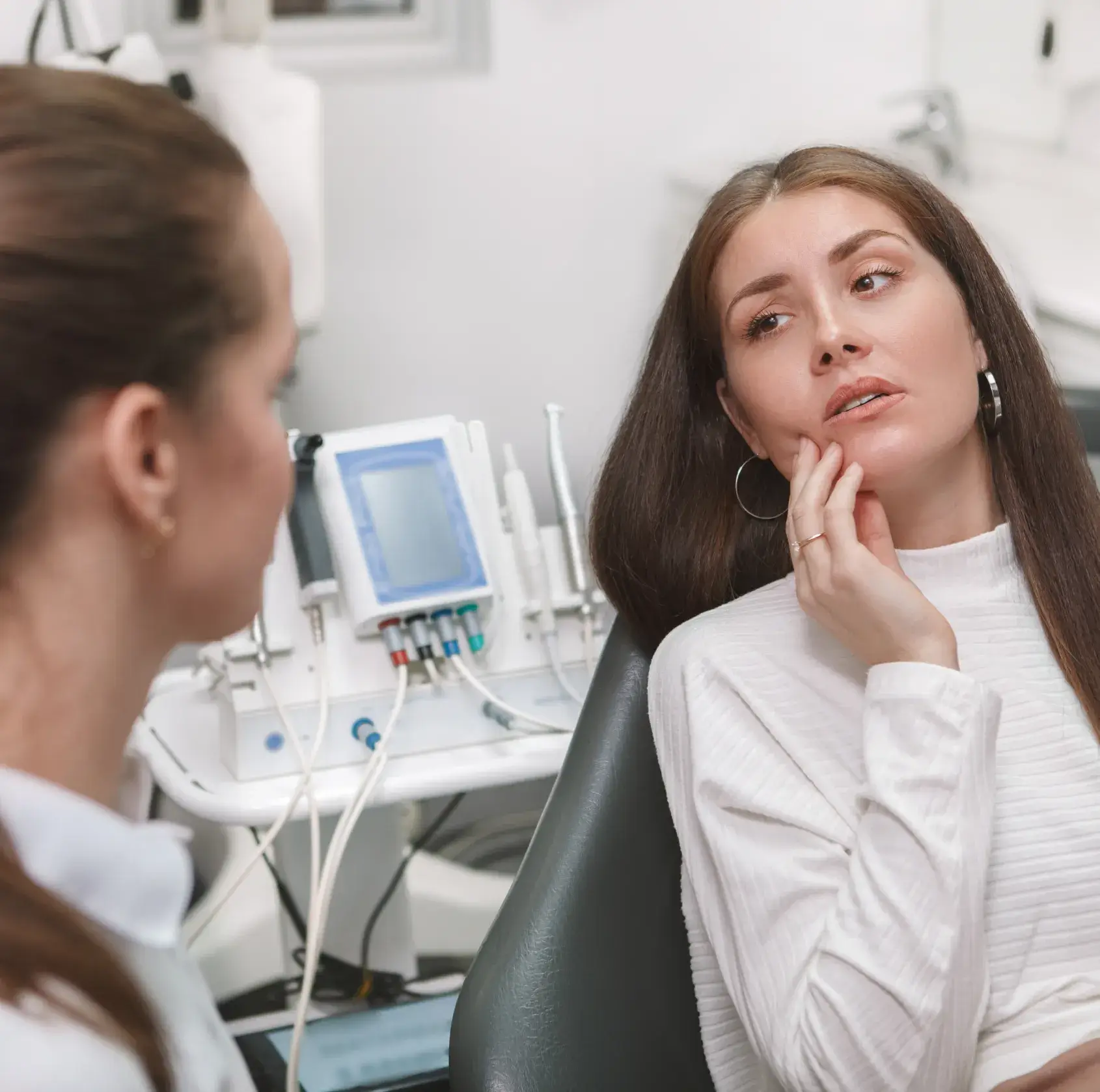 Woman in a dental office holding her cheek in pain while talking to a dentist.