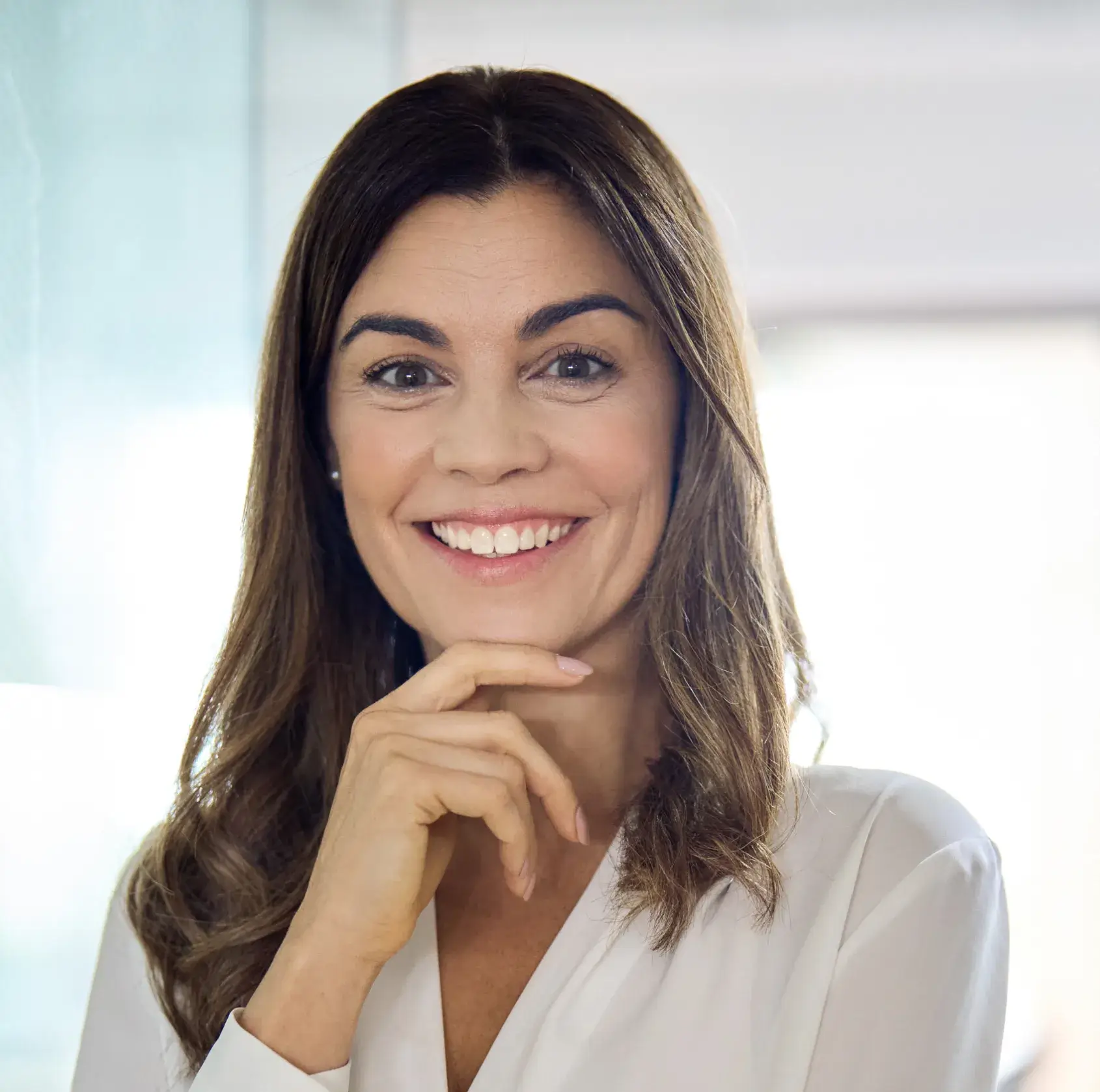 Smiling woman with long brown hair wearing a white blouse, resting her chin on her hand.