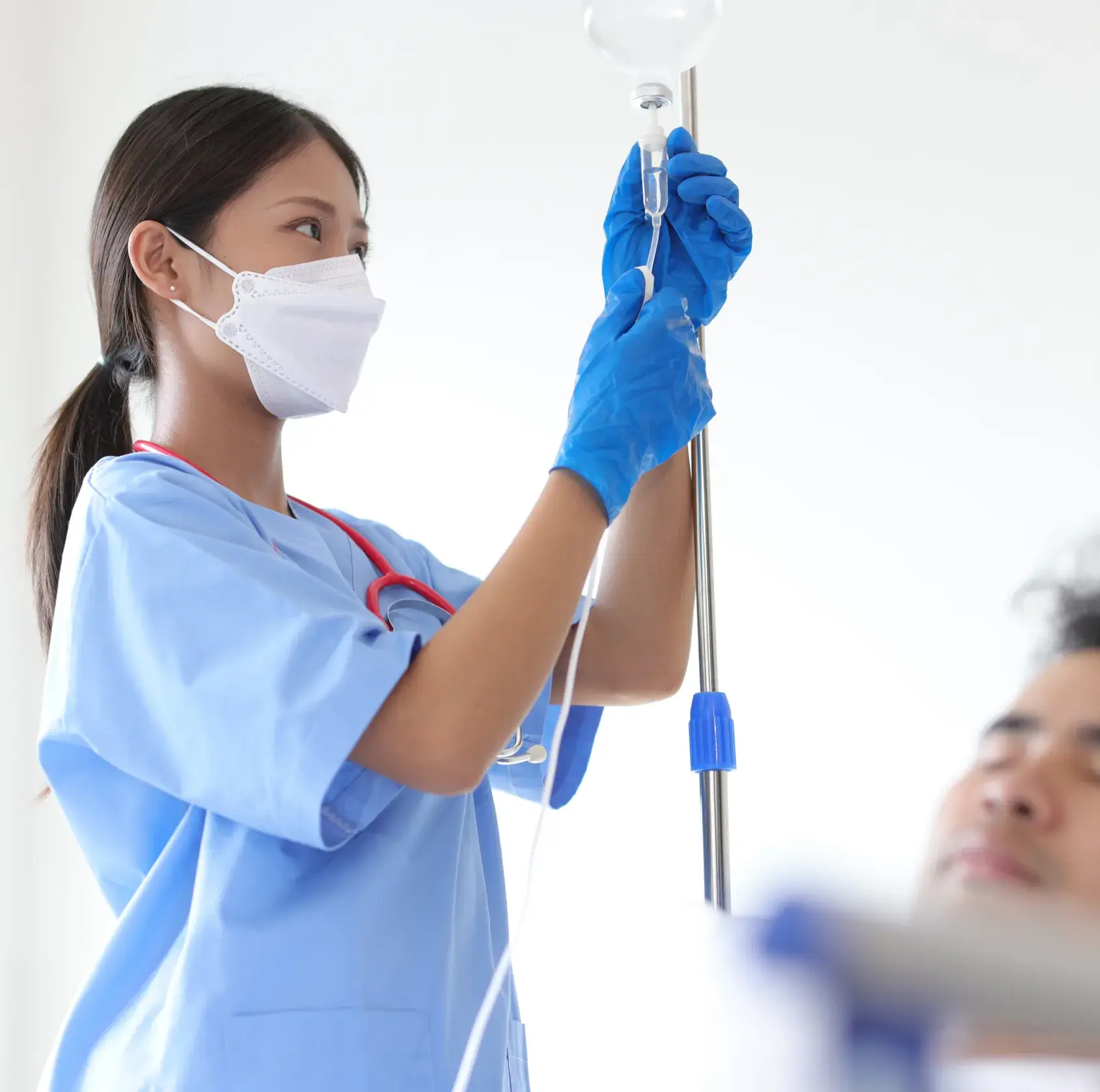 Nurse wearing blue scrubs, gloves, and a face mask adjusts an IV drip for a patient lying in bed.